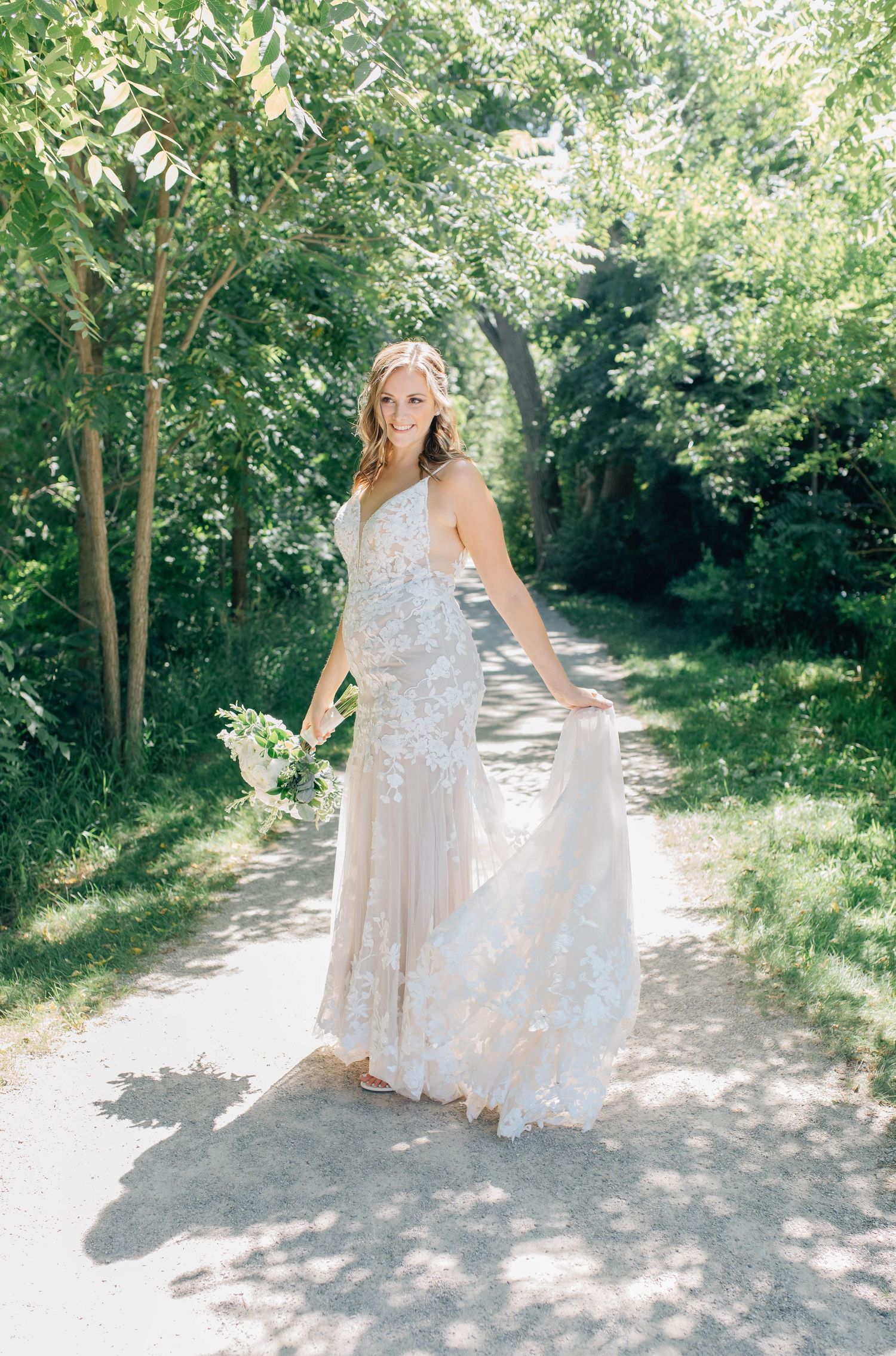 Smiling bride in floral lace gown twirling on a sunny tree-lined path.