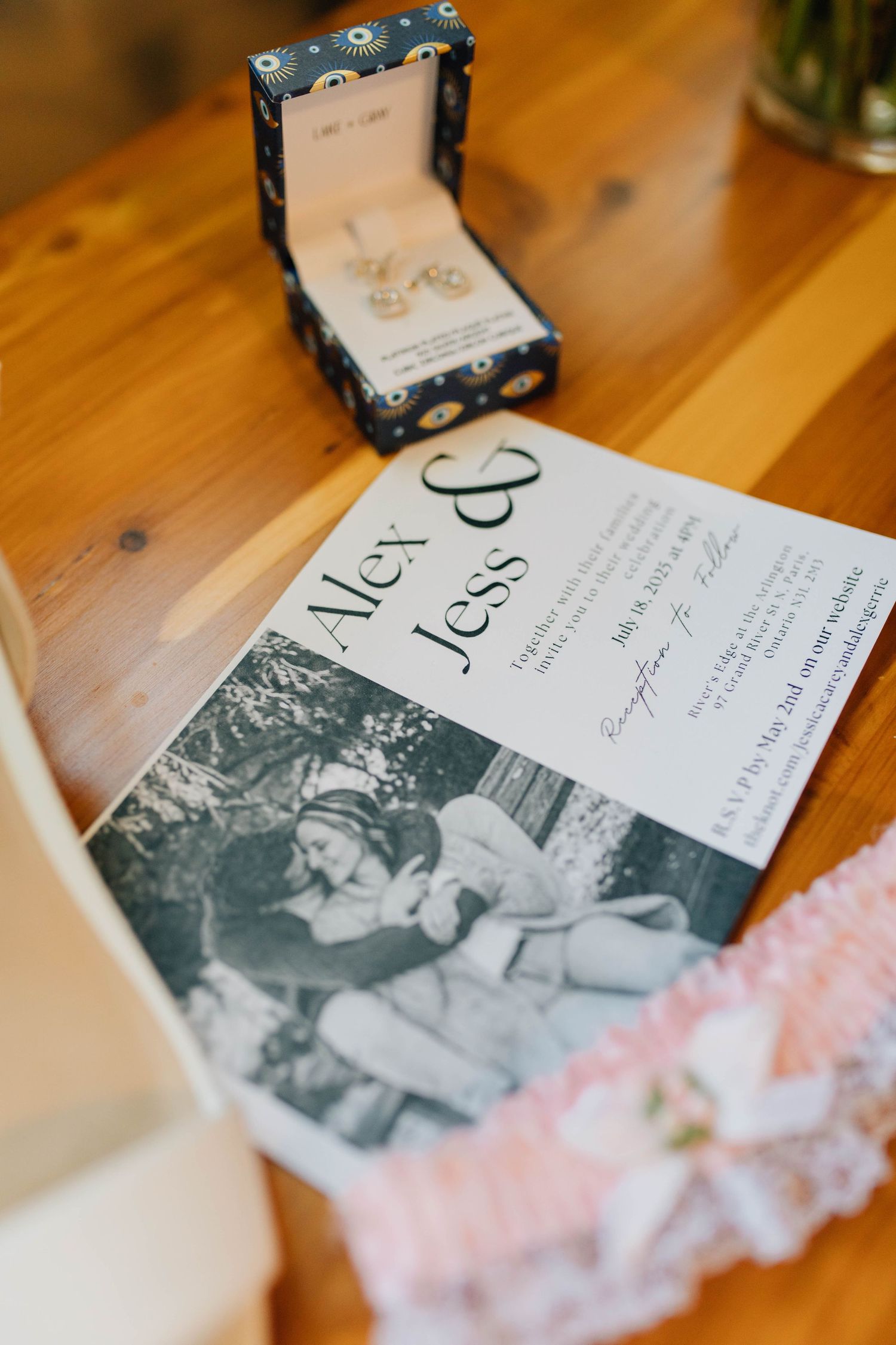 Wedding invitation for Alex and Jess displayed on wooden table with jewelry box and pink lace garter.