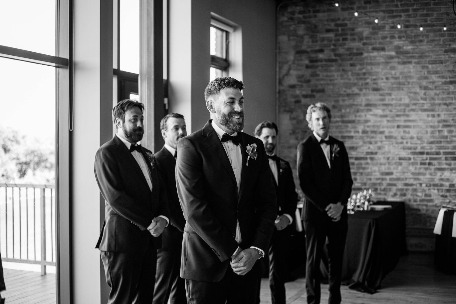 Black and white photo of emotional groom in tuxedo watching bride walk down aisle with groomsmen.