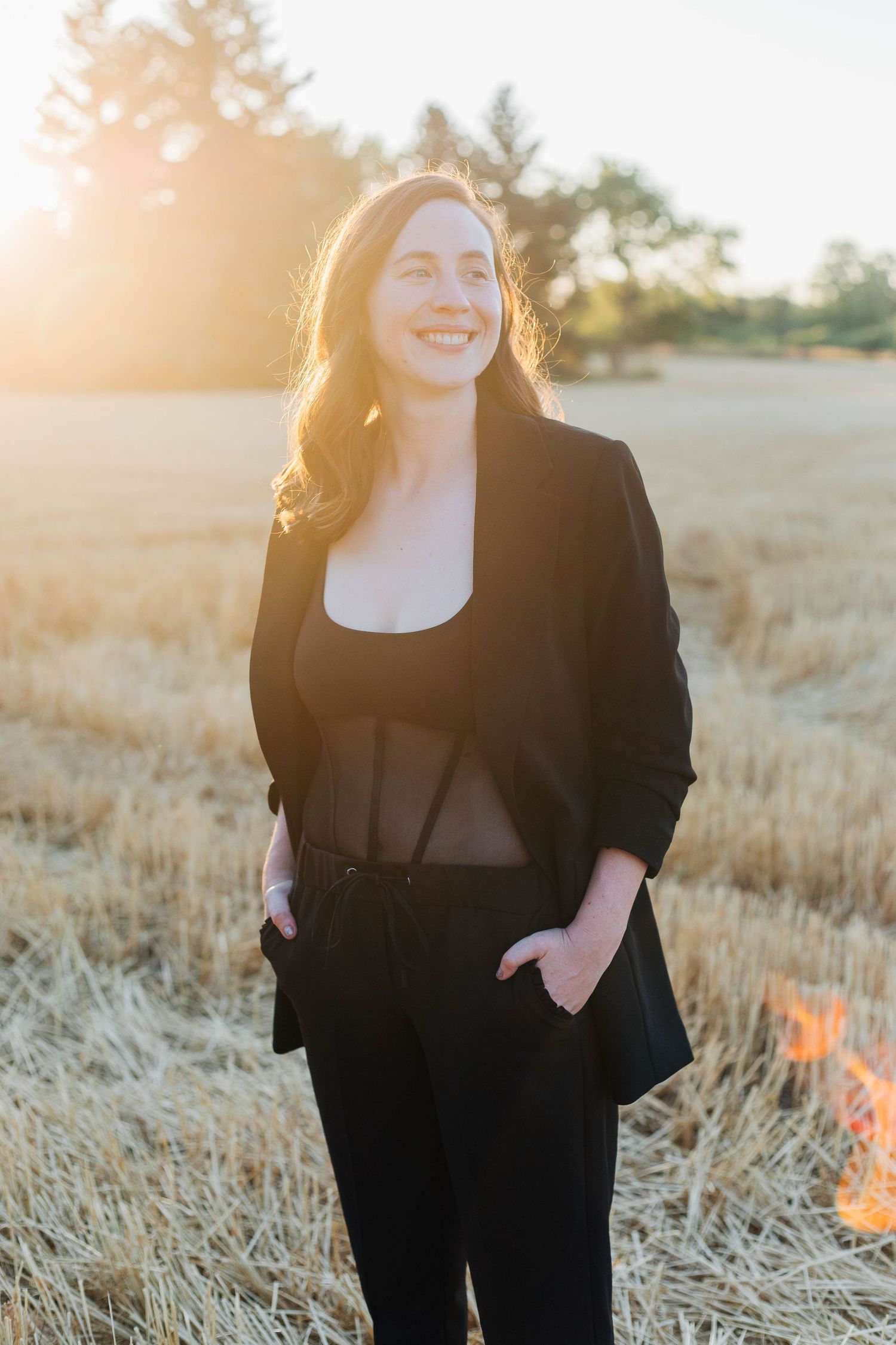 Smiling woman in black blazer and pants standing in harvested field bathed in warm golden sunset light.