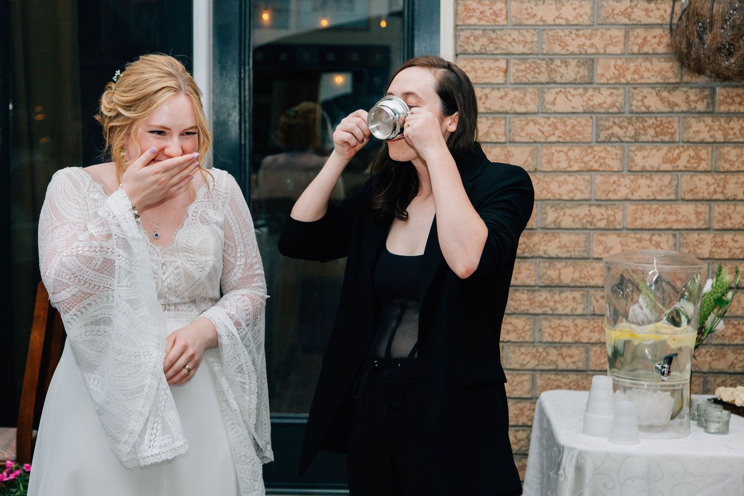 Bride laughing while wedding guest drinks from a cup at an outdoor wedding reception.