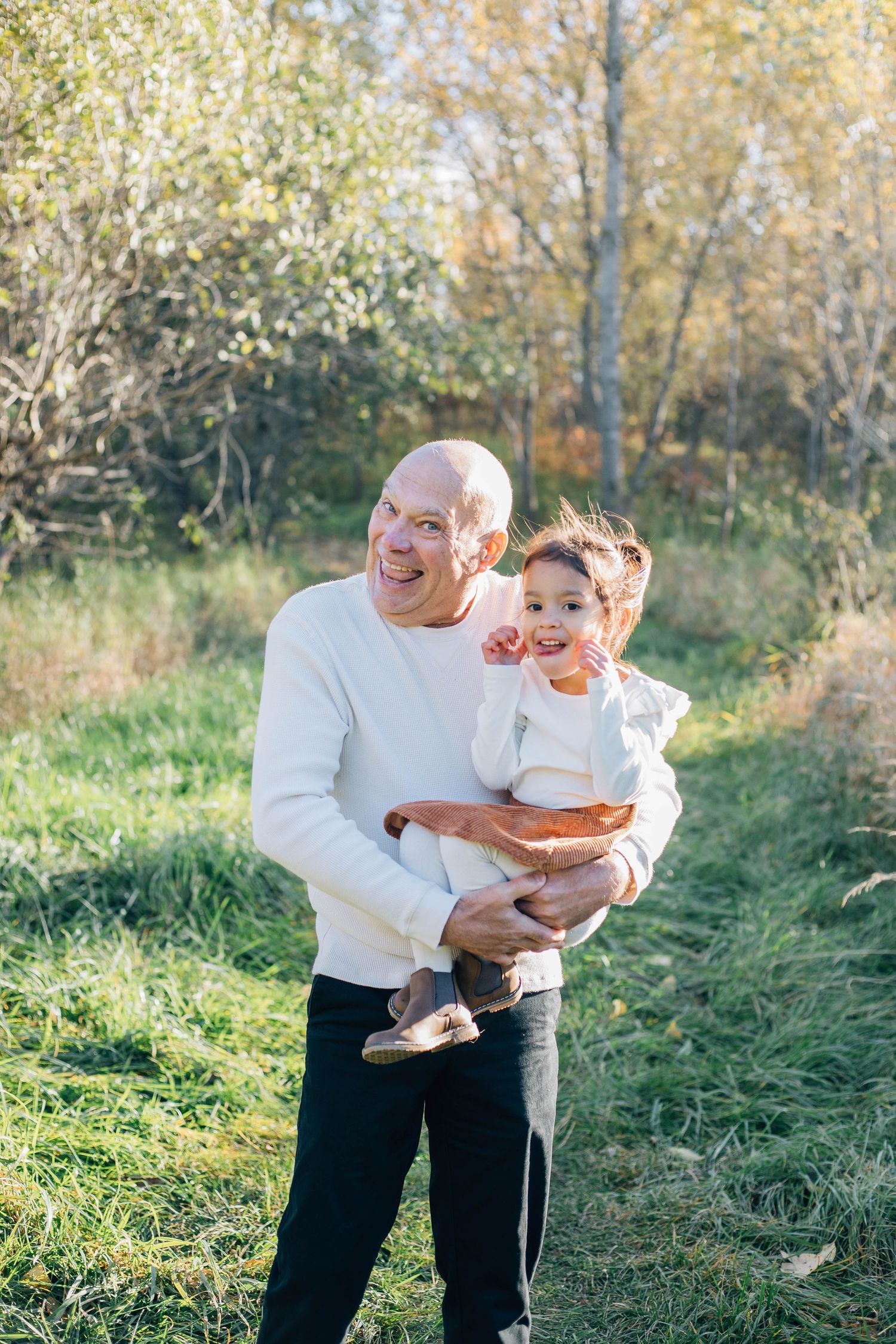 Bald man in white sweater laughing while holding a smiling toddler outdoors in autumn setting.