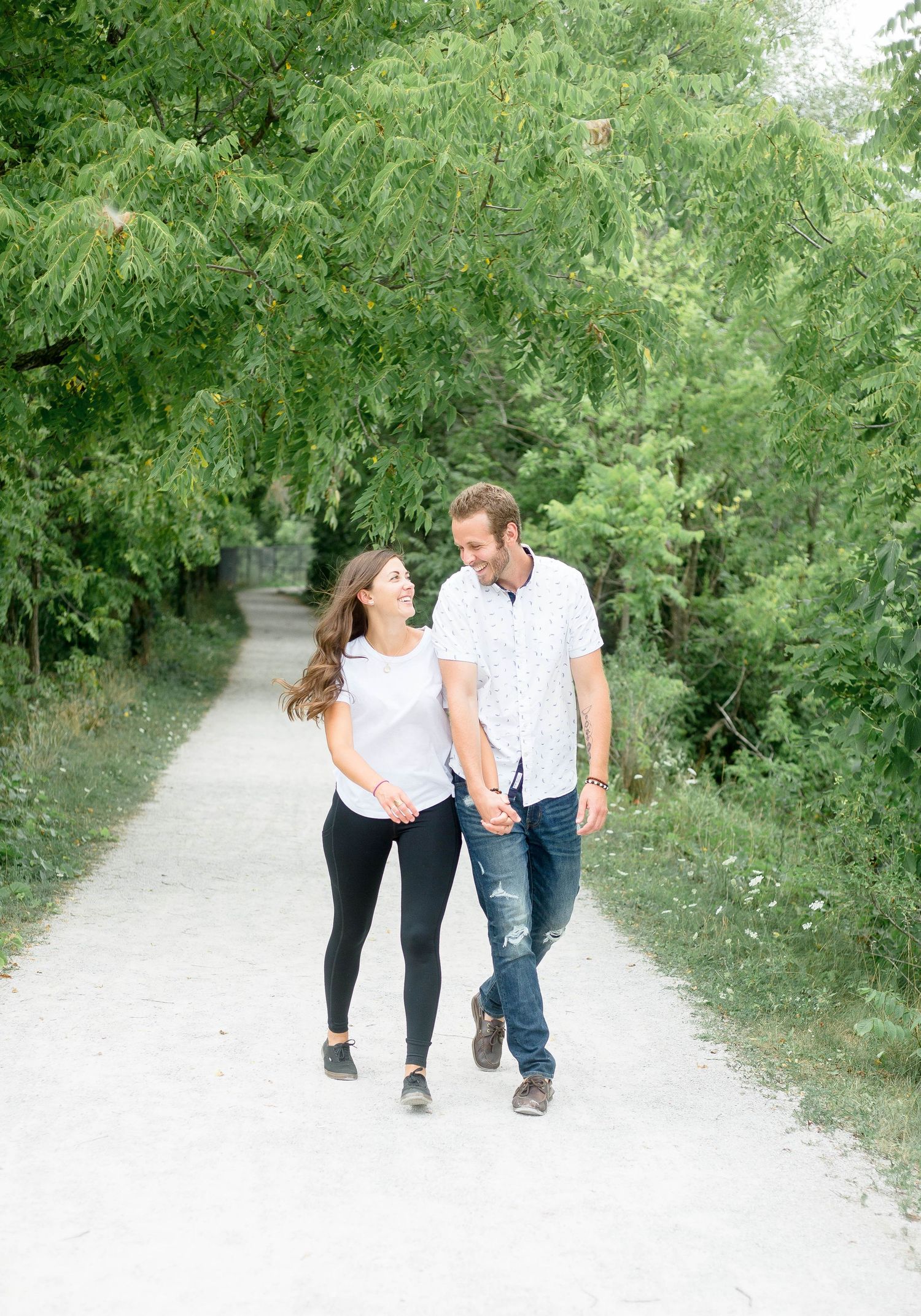 A happy couple holds hands while walking along a bright tree-lined gravel path outdoors.