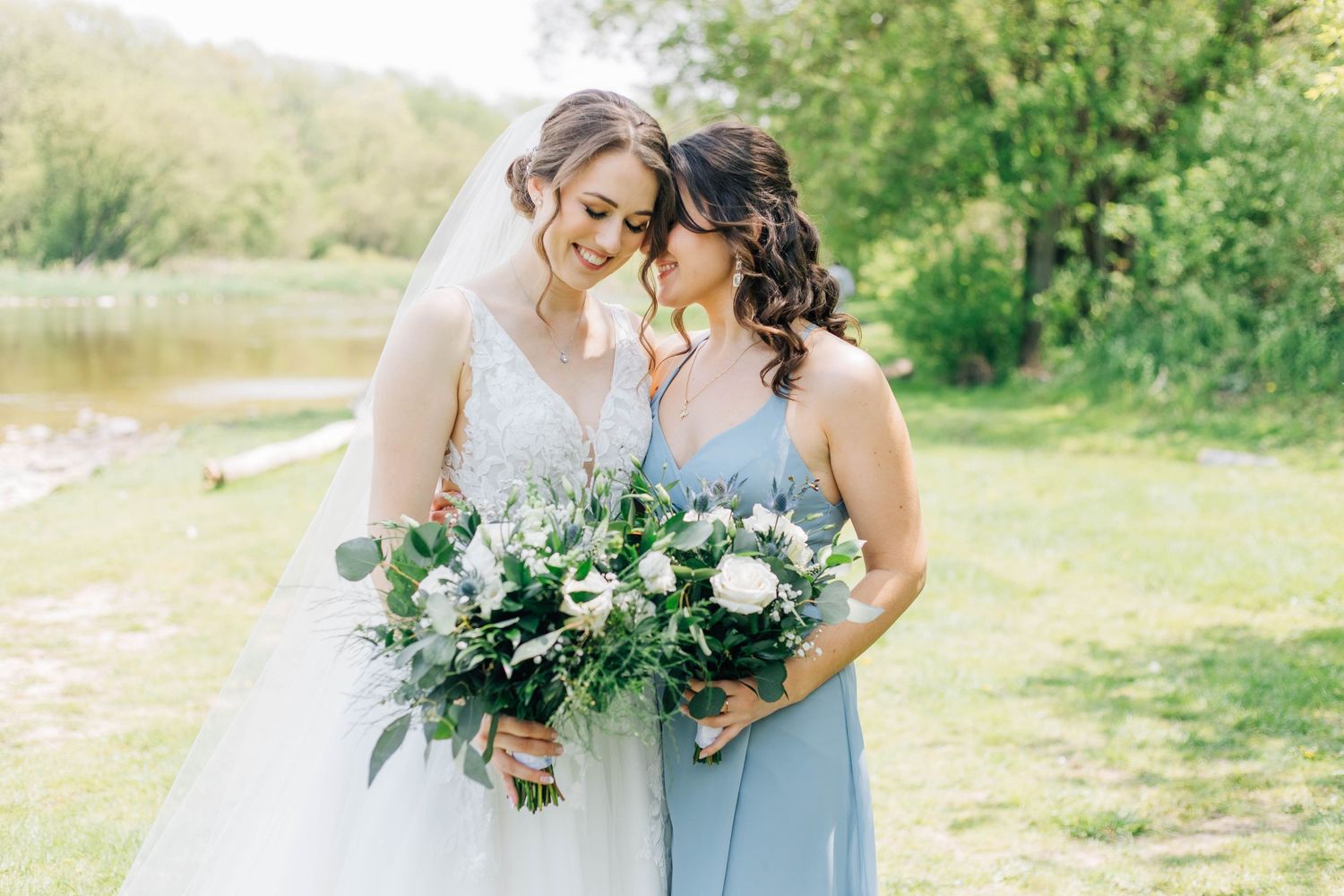 Bride in lace gown and bridesmaid in blue dress share a tender moment holding white floral bouquets.