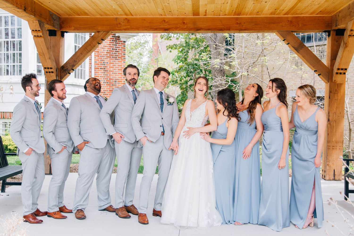 Joyful wedding party laughs together under wooden pavilion with bridesmaids in dusty blue gowns.