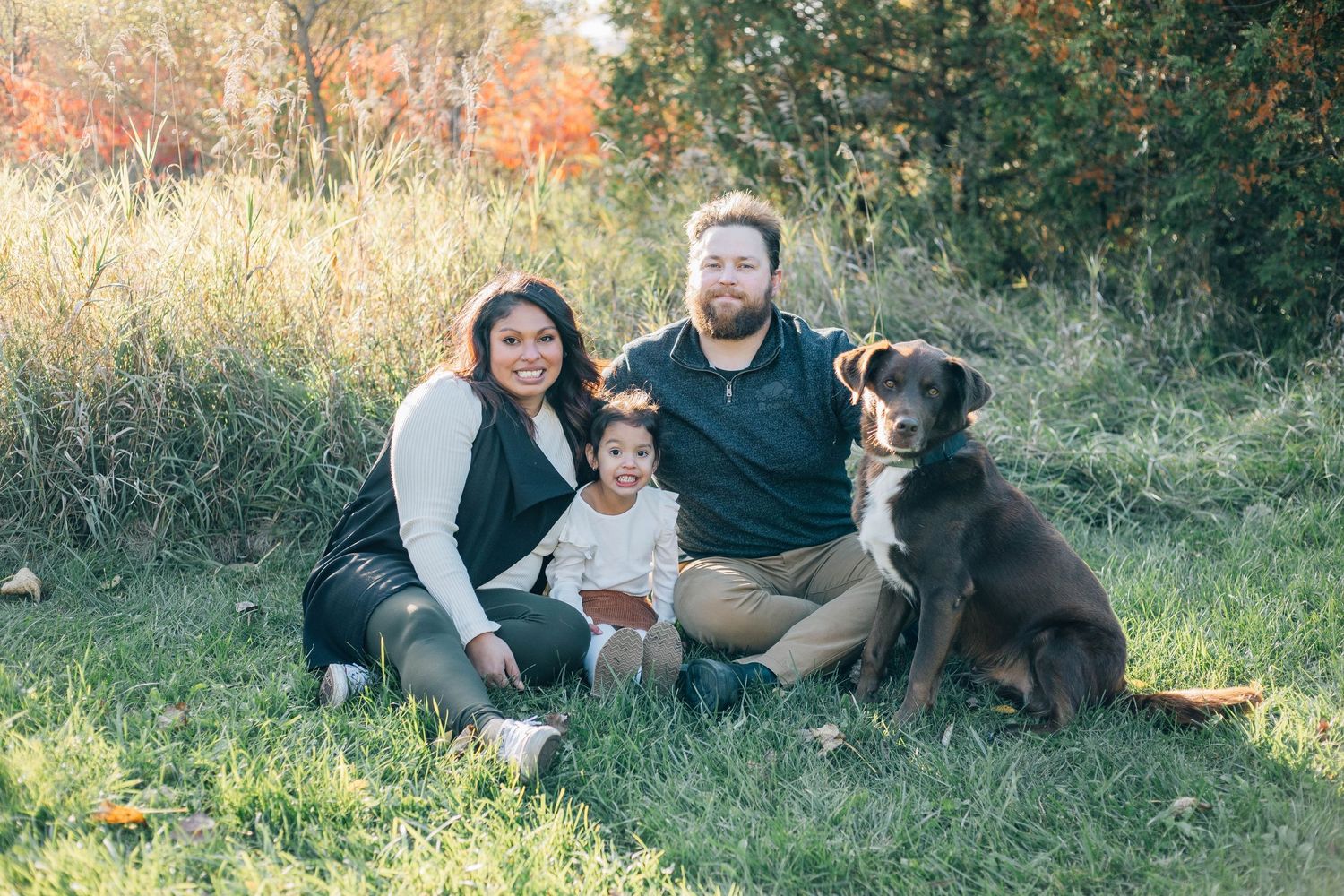 Family with young child and chocolate Lab dog sitting together in autumn grass outdoors.