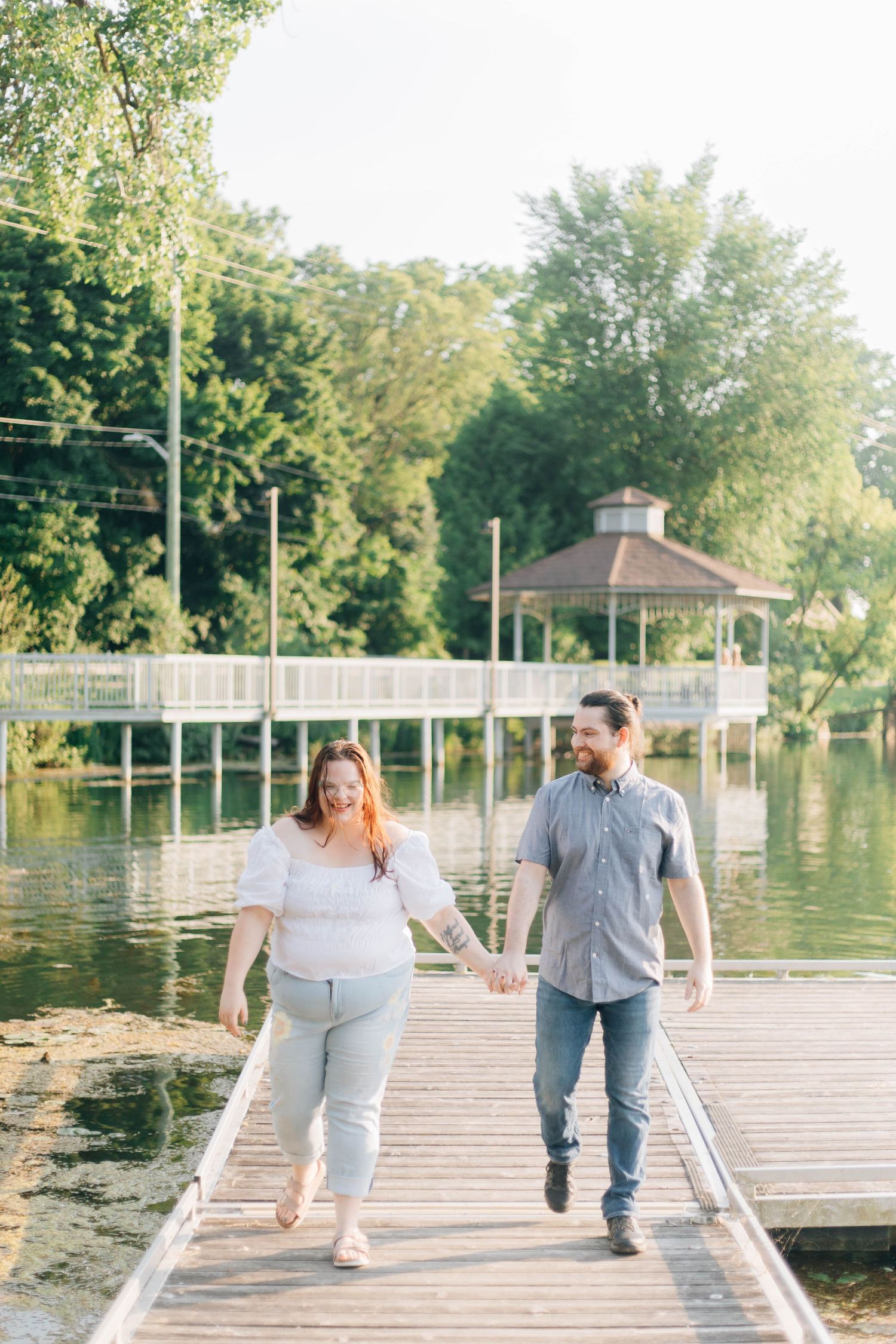 Couple holding hands and walking along a wooden dock with a gazebo and lake in background.