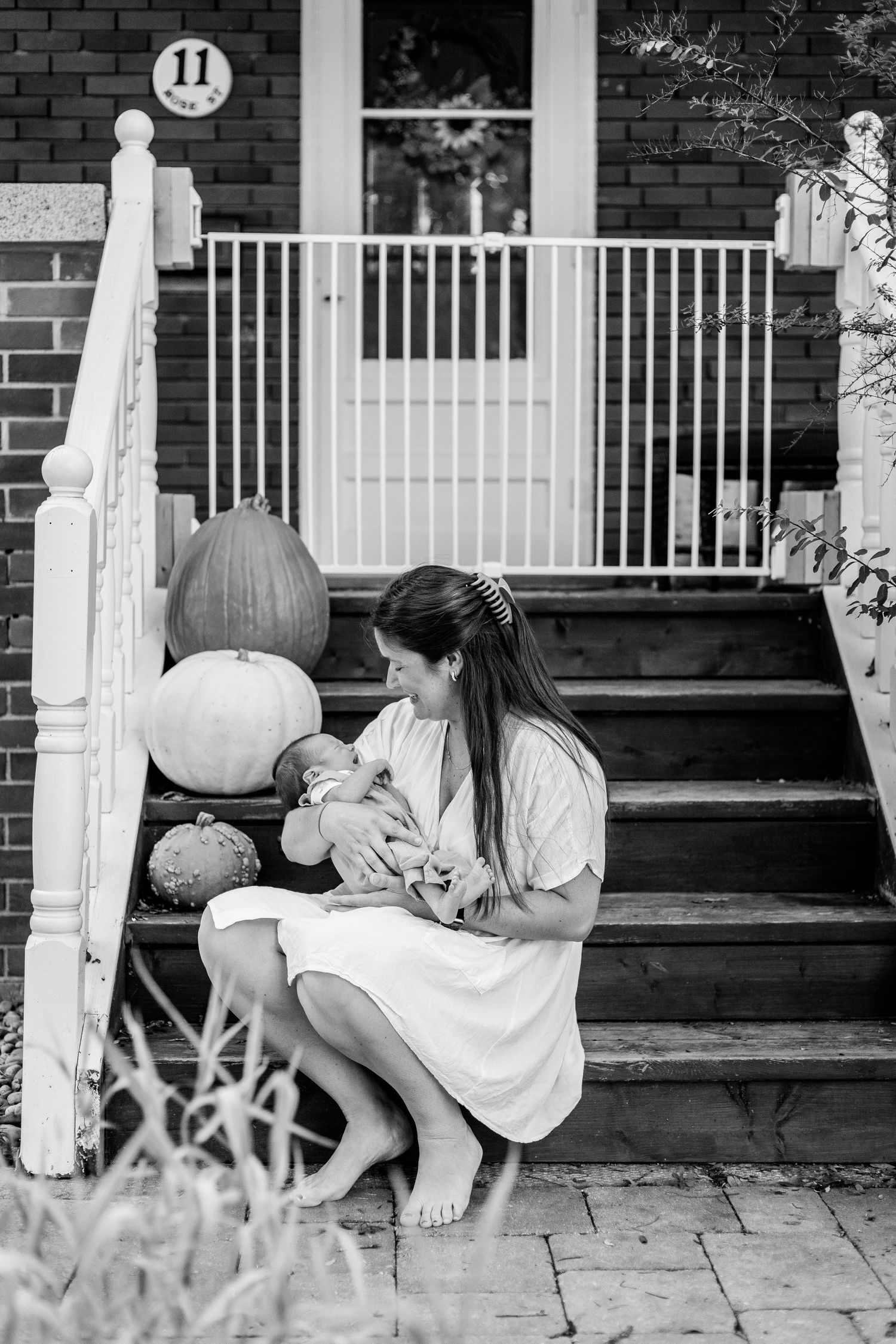 Black and white photo of barefoot mother sitting on front porch steps holding newborn baby with pumpkins beside her.