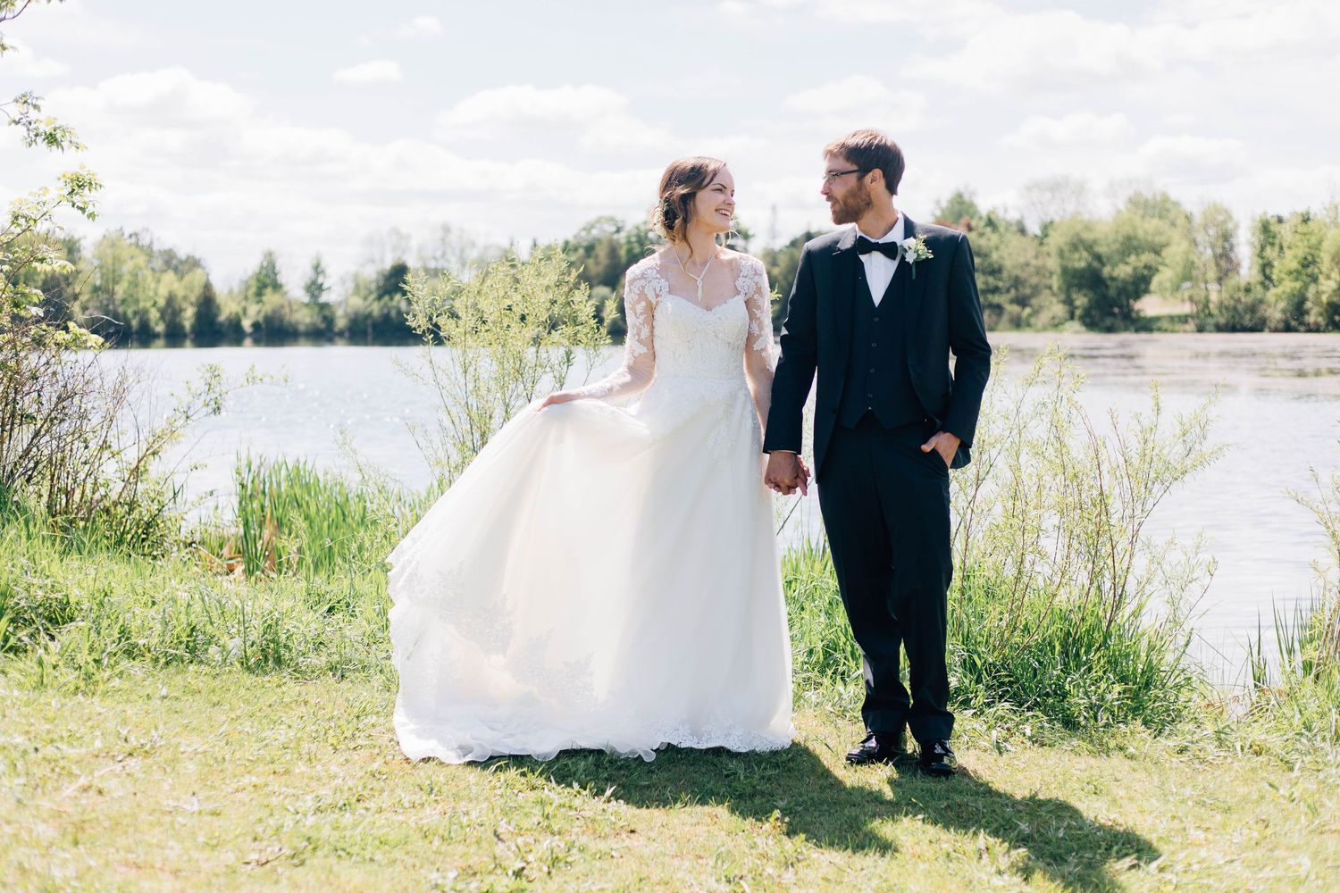 Bride and groom walking hand in hand along a sunny lakeside, bride in elegant lace wedding gown.