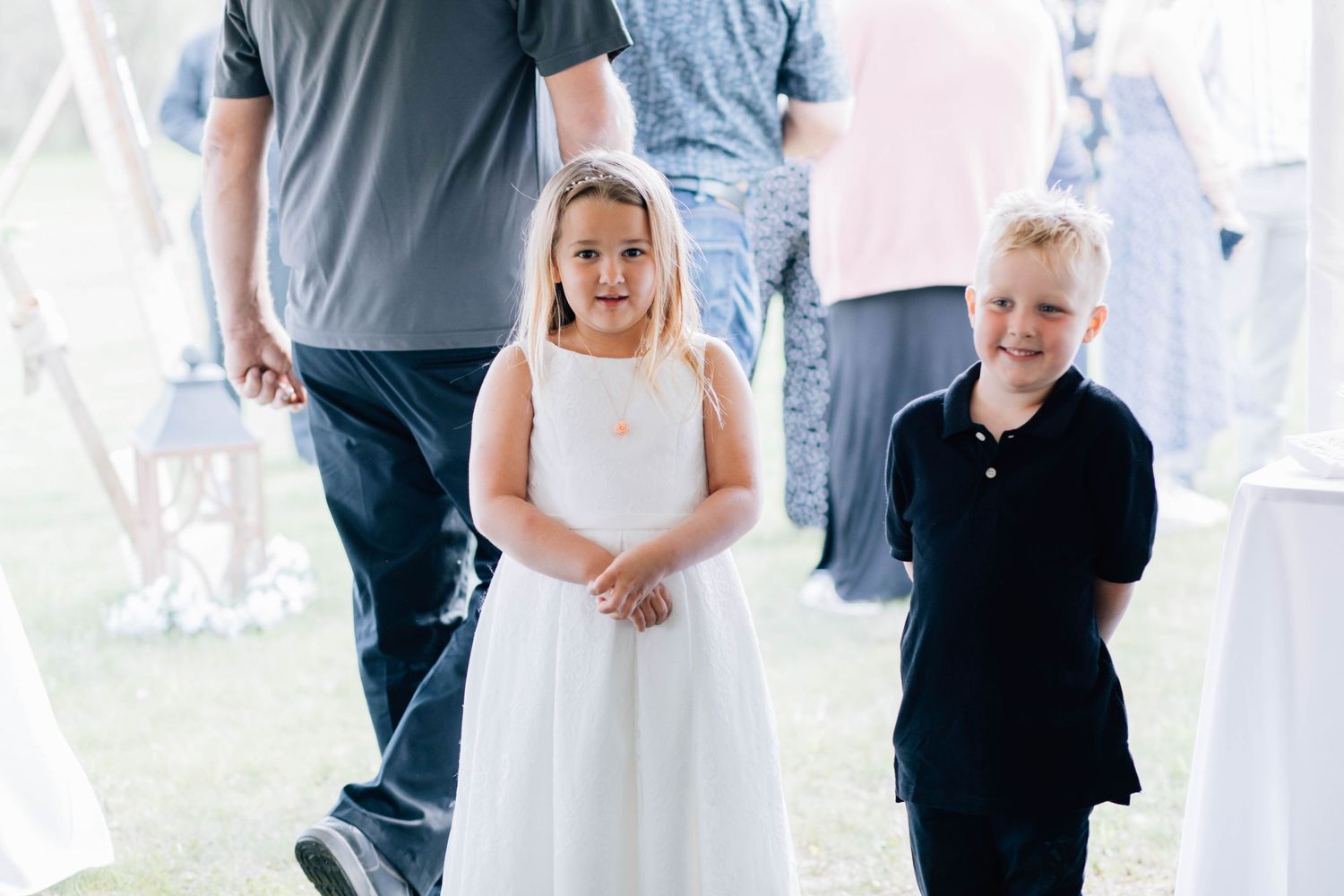 Two young children, a girl in white dress and boy in navy, walking at an outdoor wedding celebration.