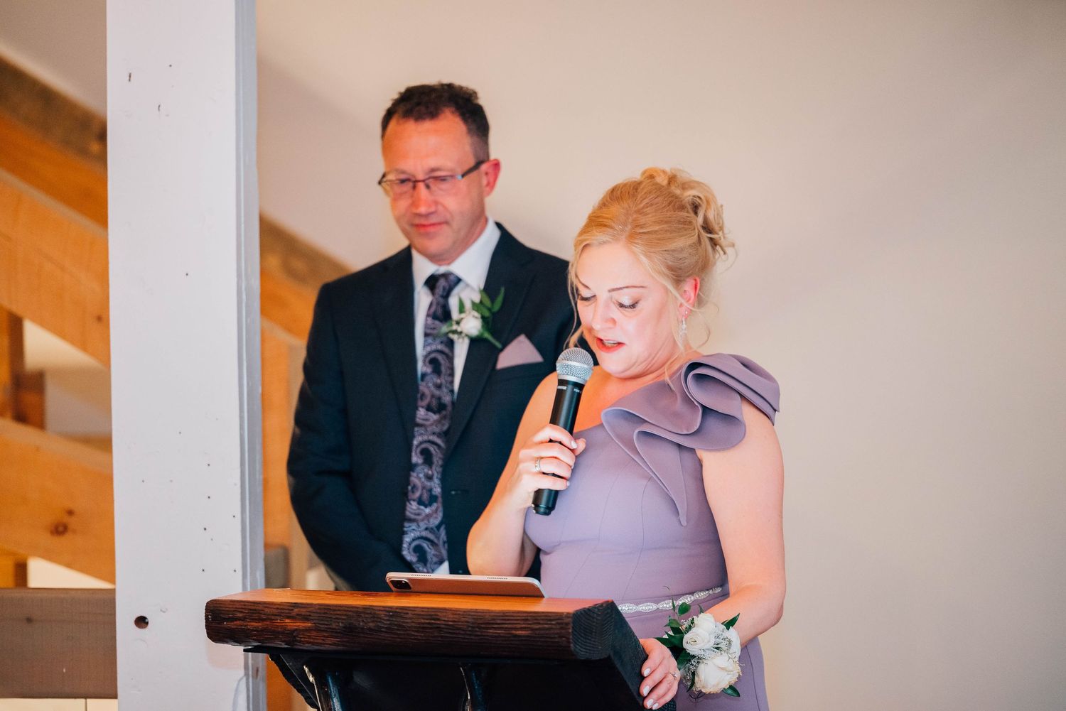 Bridesmaid in lavender dress giving wedding speech at podium with man standing beside her.