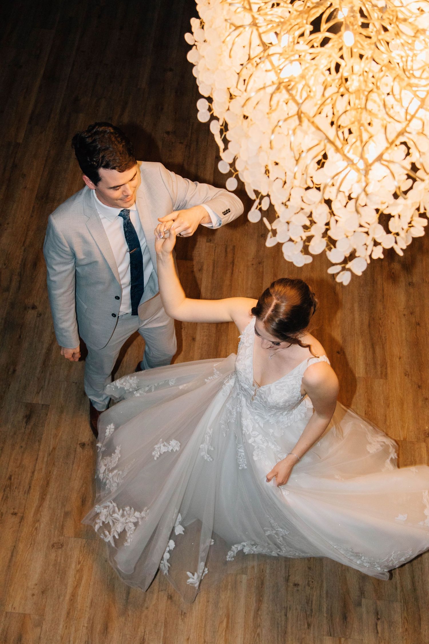 Groom spinning bride in stunning lace wedding gown beneath decorative chandelier on dance floor.