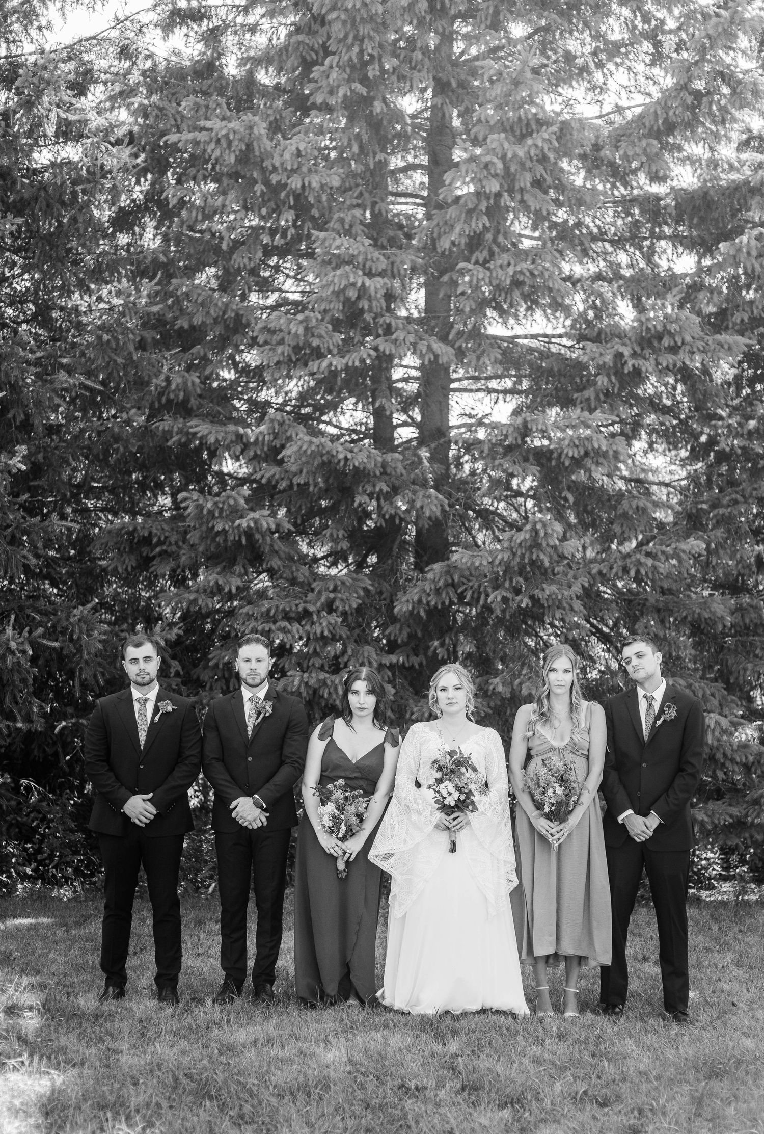 Black and white photo of wedding party of five standing solemnly before a large evergreen tree.