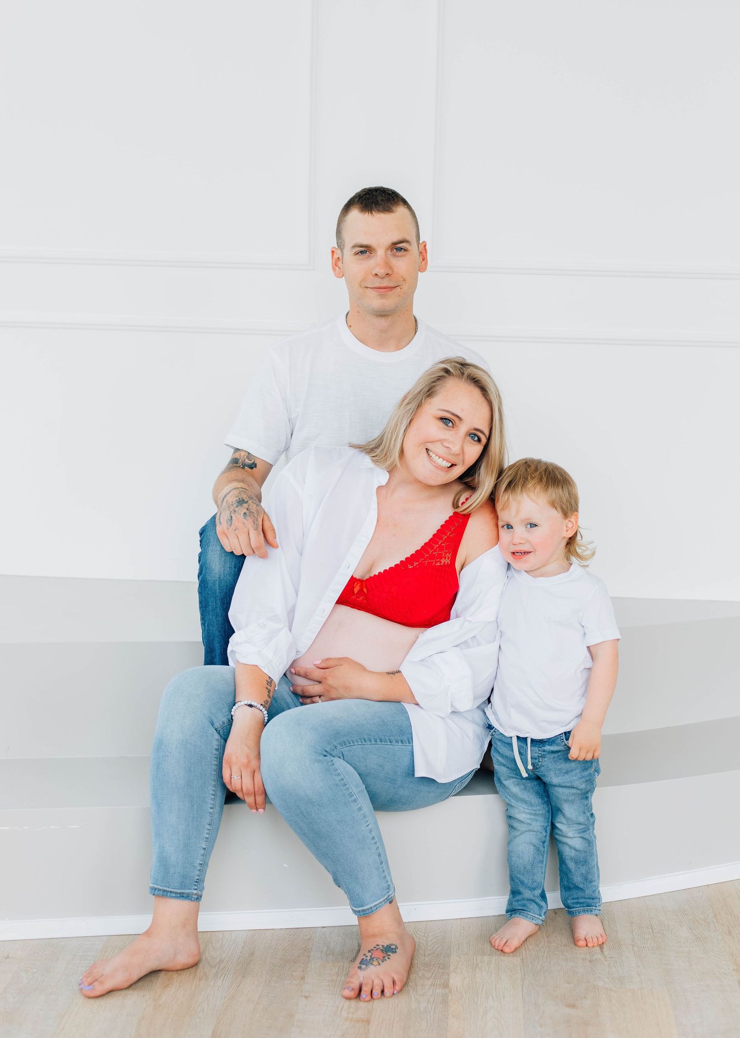 Family maternity portrait with pregnant mom in red bralette seated between tattooed partner and young toddler.