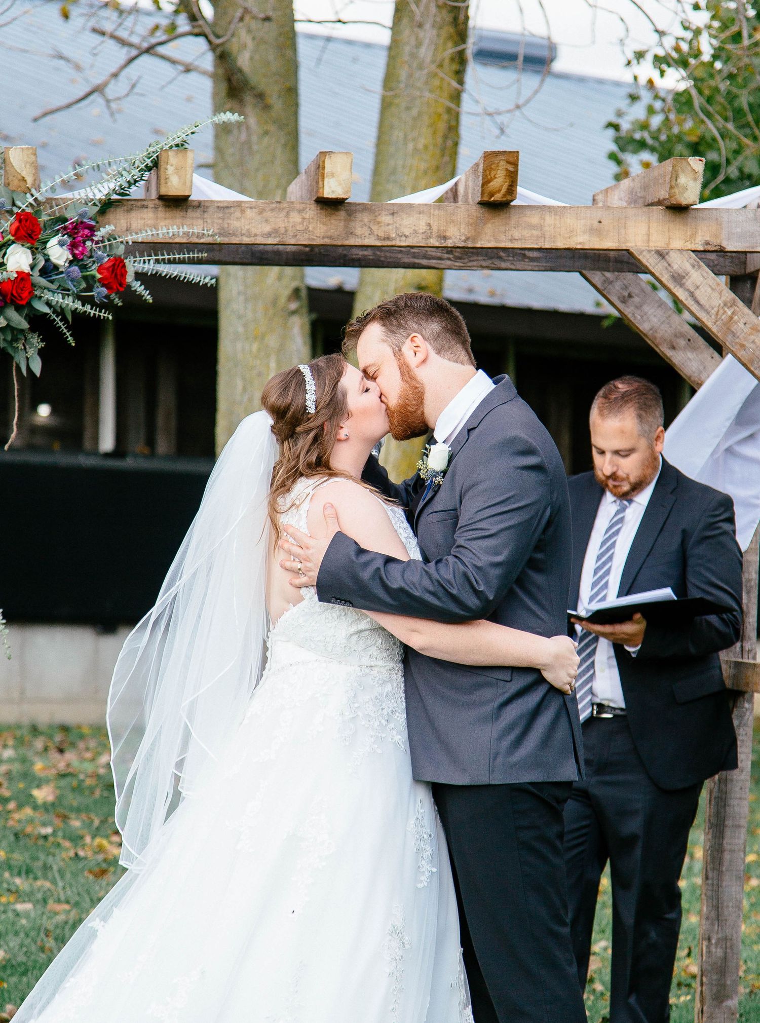 Bride and groom share first kiss at outdoor wedding ceremony under rustic wooden arch with floral decorations.