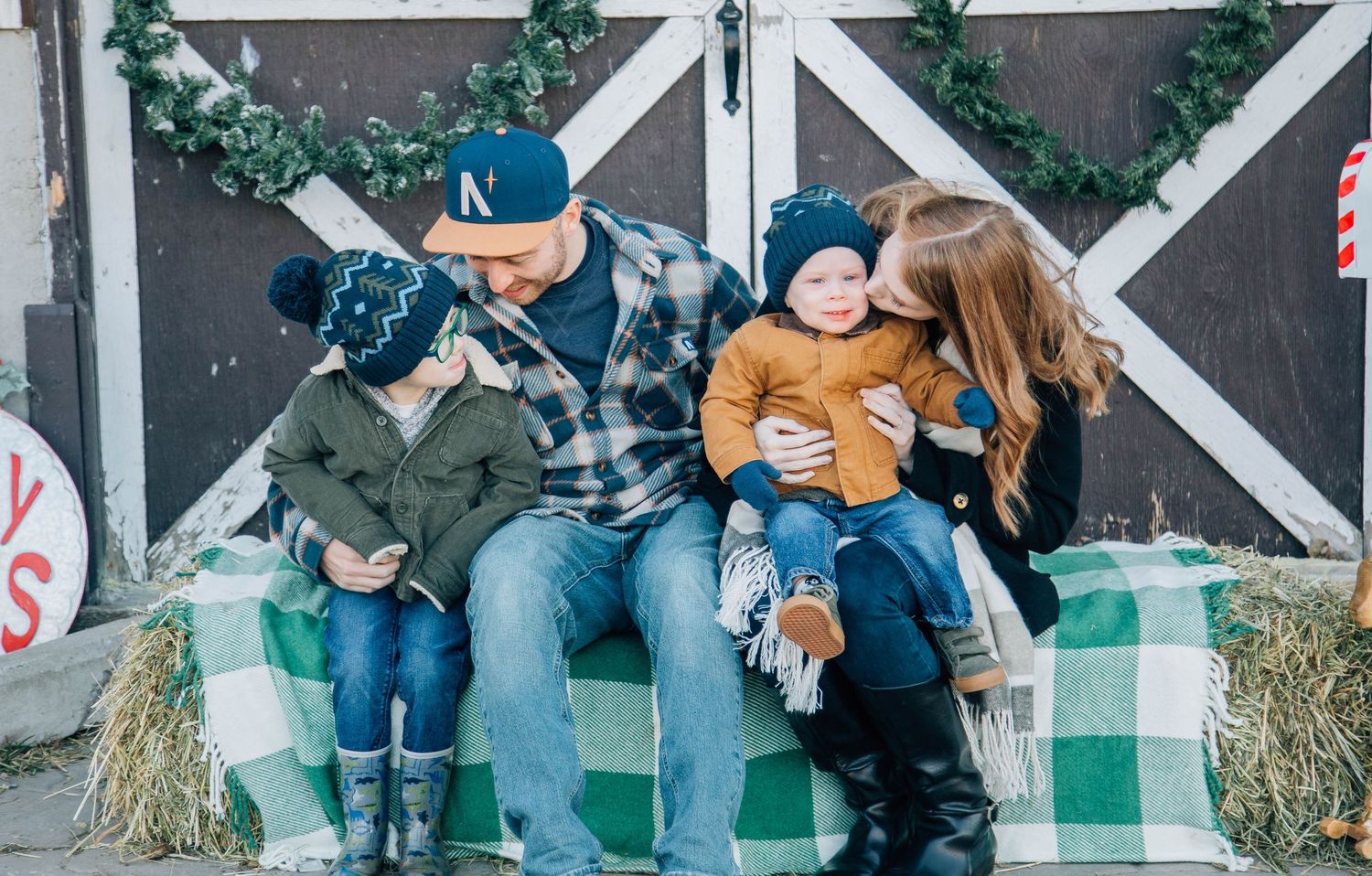 Family of four in cozy winter outfits sitting together on hay bales in front of a barn.