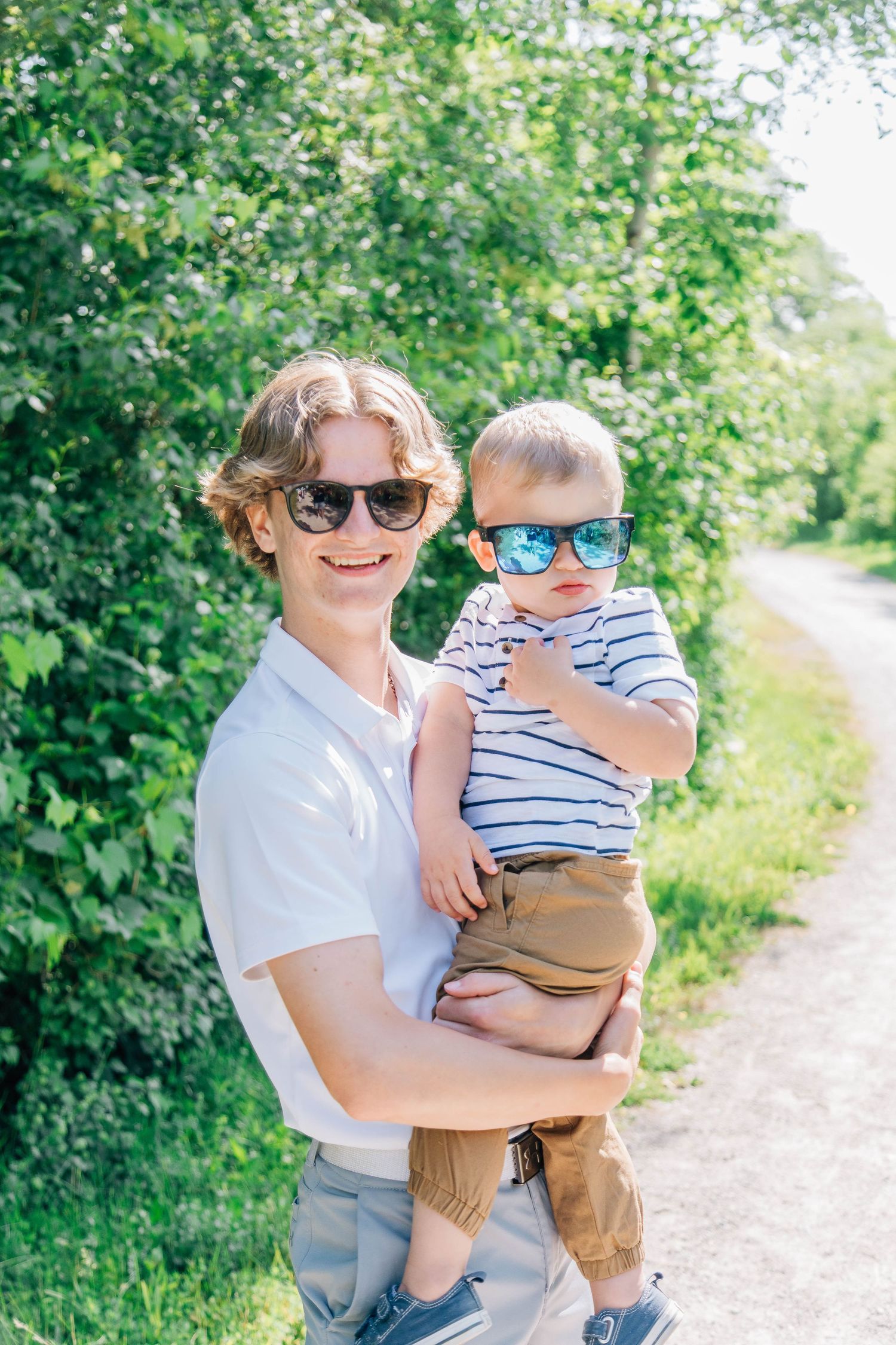 Young man smiling and holding a toddler boy, both wearing sunglasses outdoors on a sunny day.
