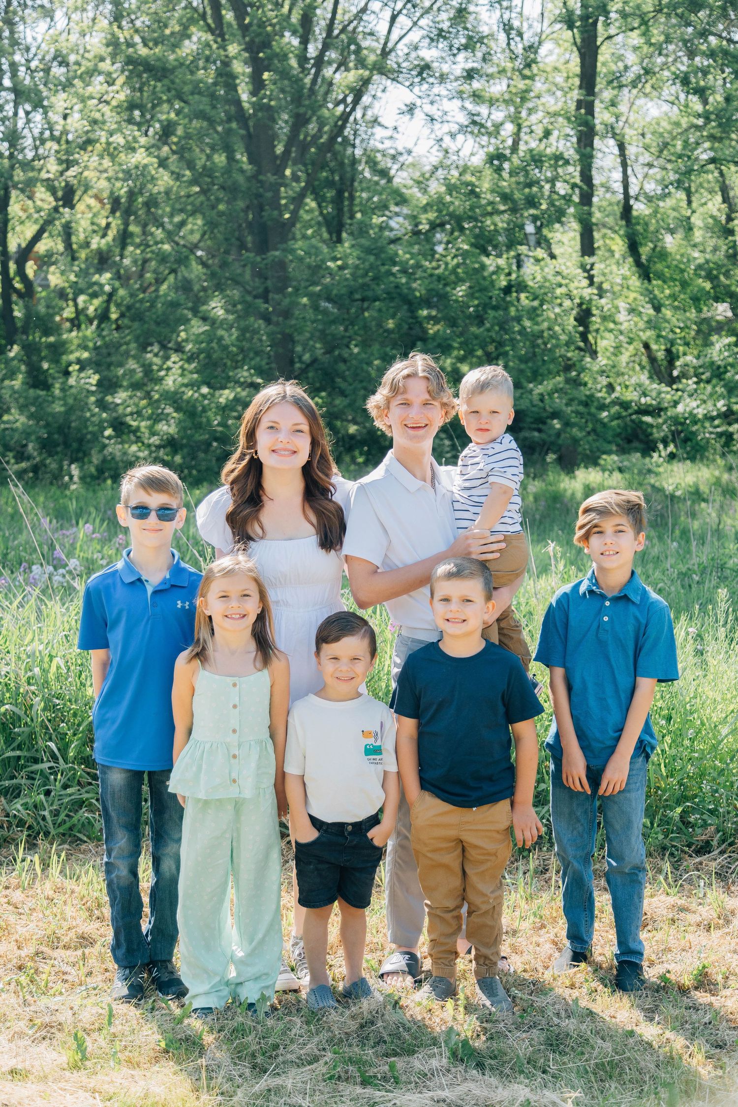 Two teenagers and six young children posing together for a group portrait in a lush green outdoor setting.