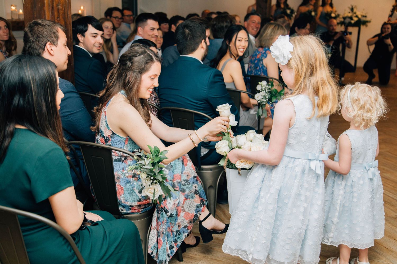 Young flower girls handing white roses to seated wedding guests indoors.
