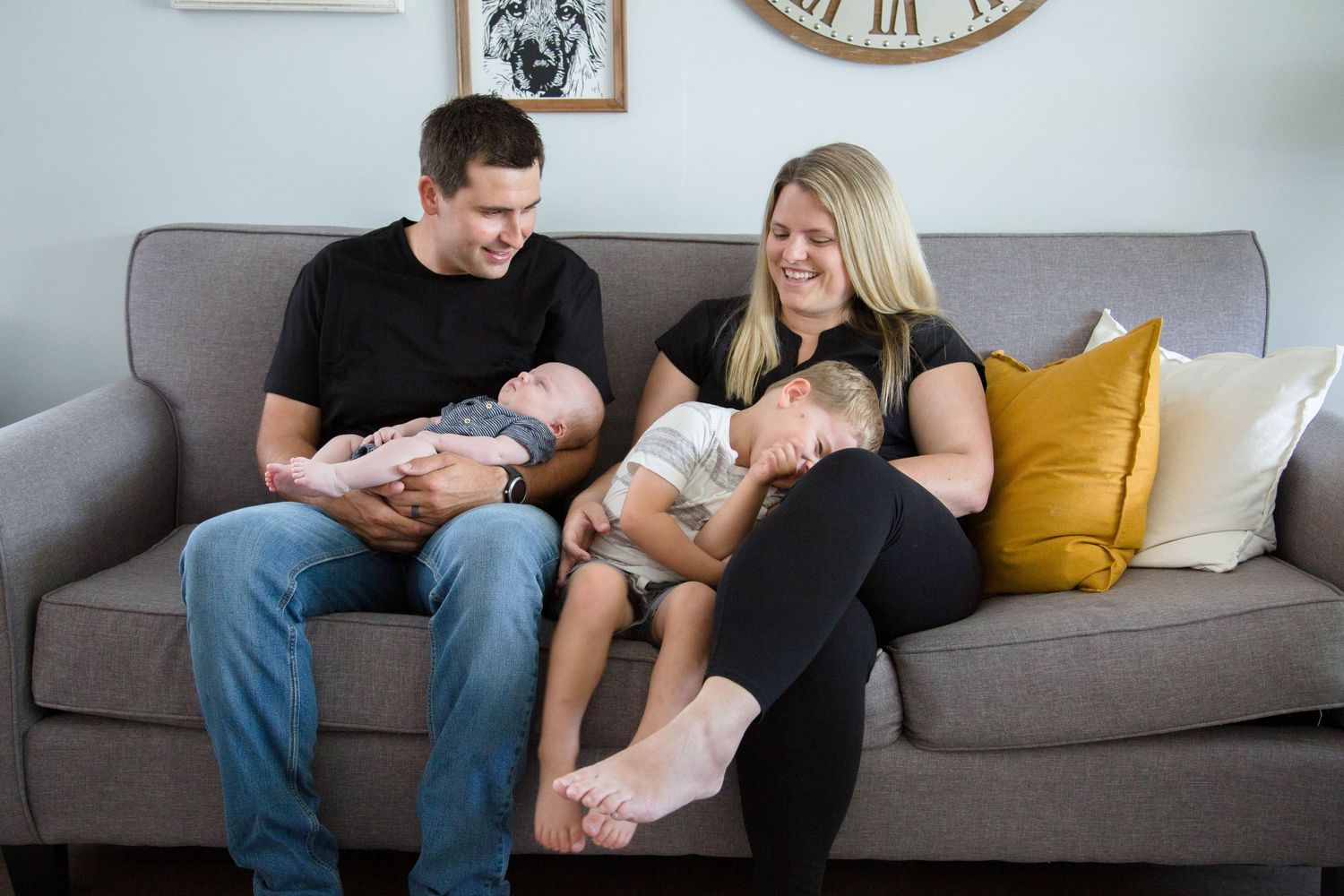 Family of four relaxing on gray sofa, parents each holding a young child in cozy home setting.