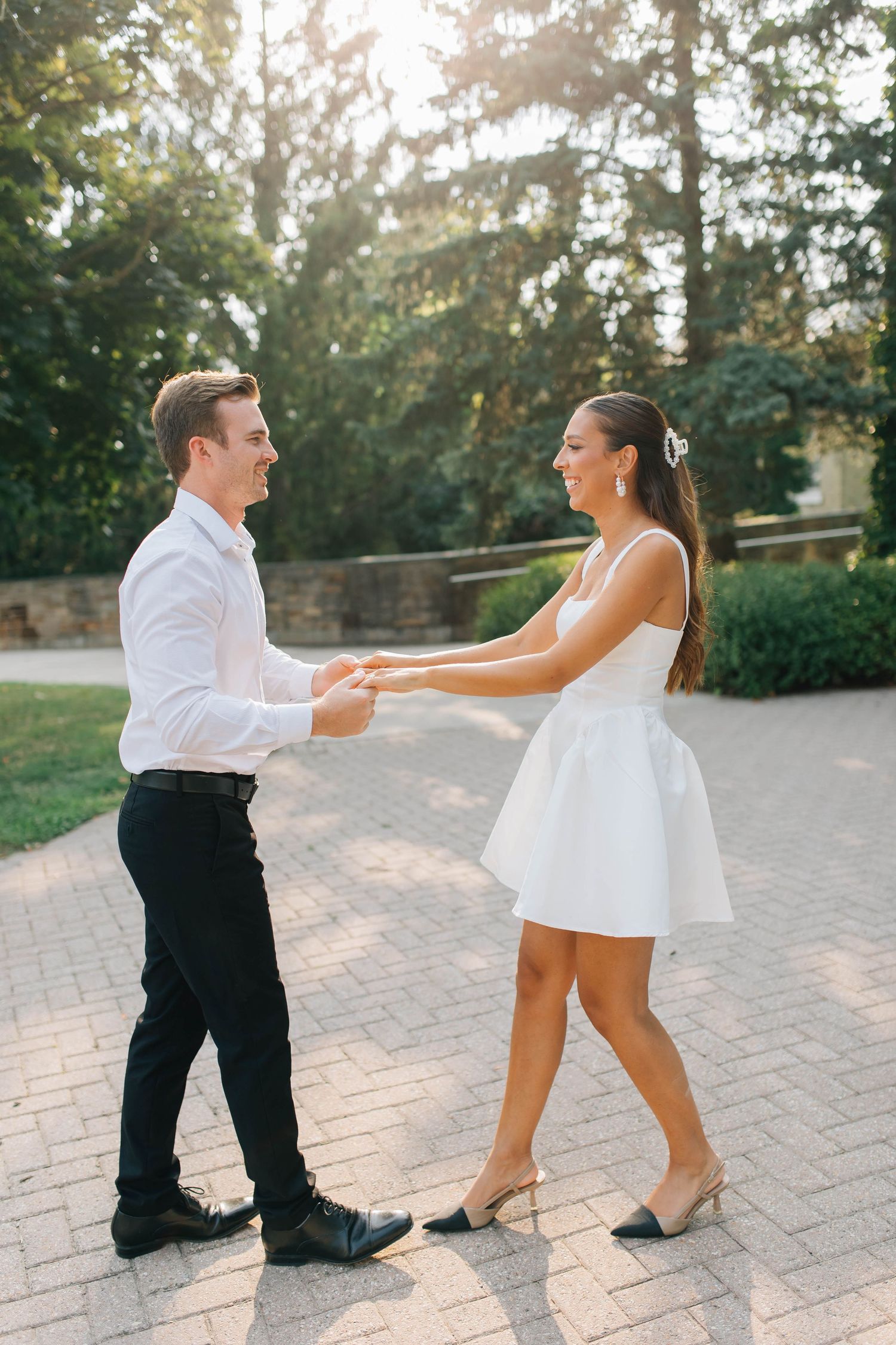 Couple holding hands and dancing outdoors on a brick path surrounded by lush green trees in golden sunlight.