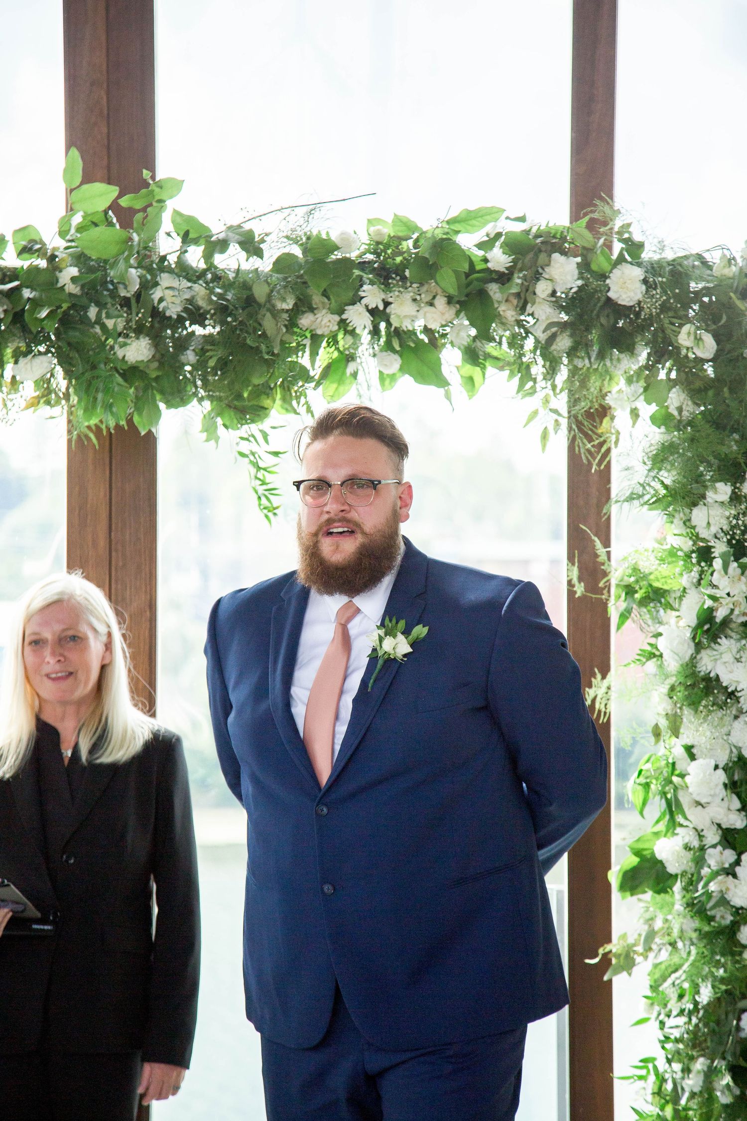 Bearded groom in navy suit with pink tie stands at floral wedding arch decorated with white flowers.