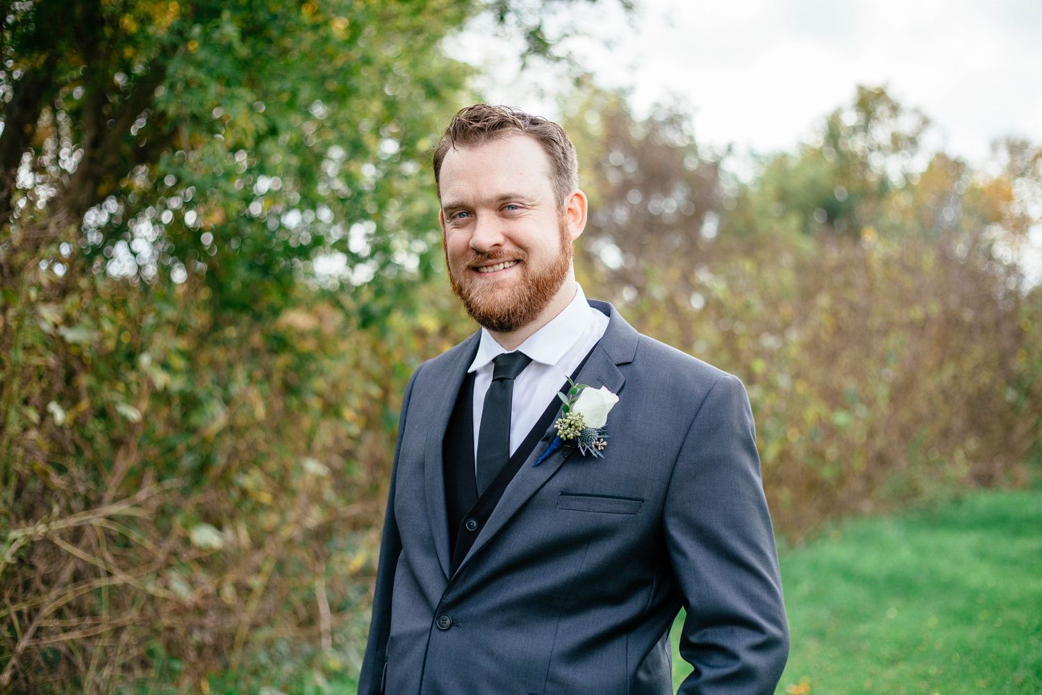 Smiling groom in gray suit with white boutonniere standing outdoors among colorful autumn foliage.