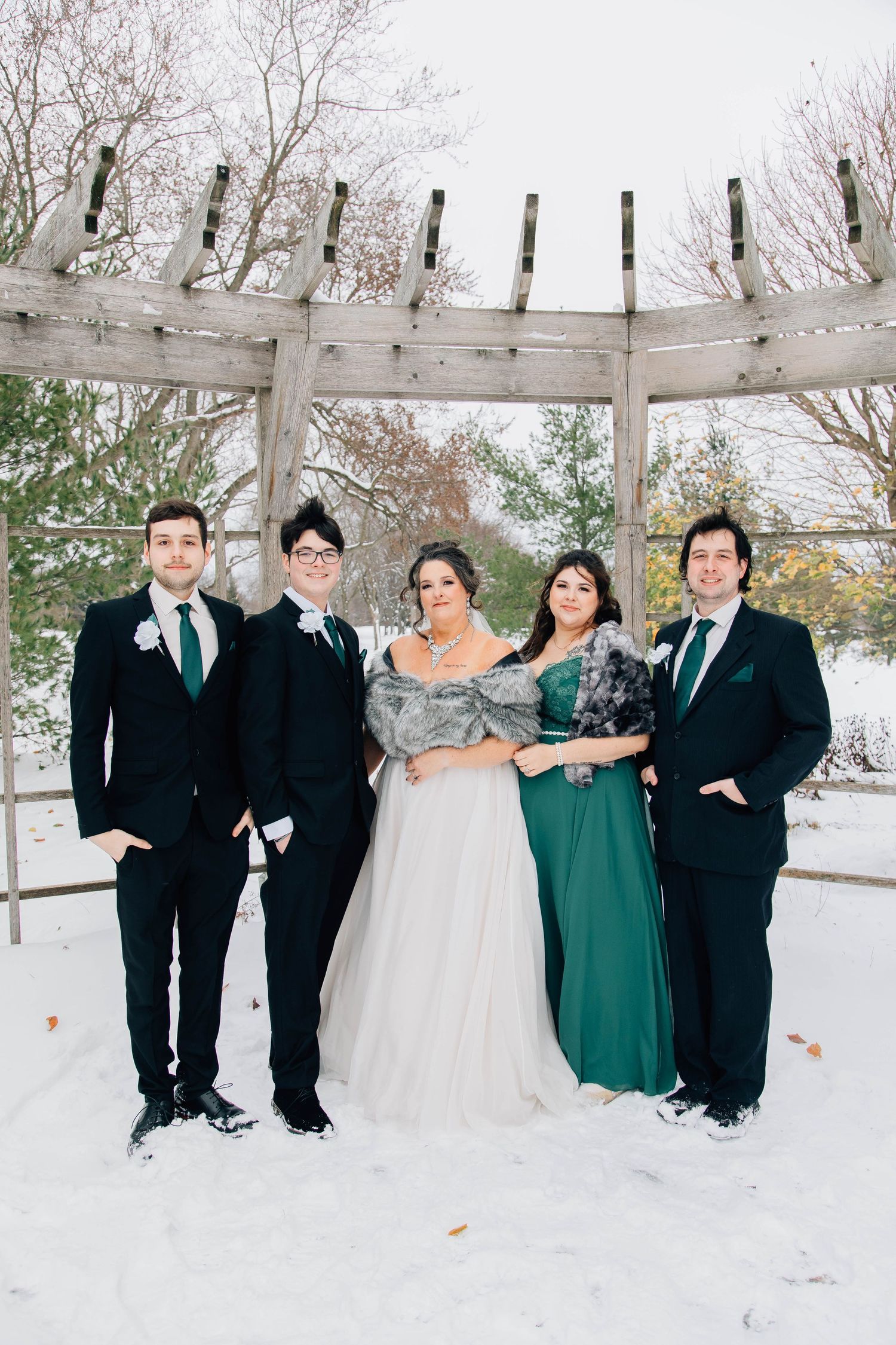 Winter wedding party poses in snow under wooden pergola in emerald green and black formal attire.
