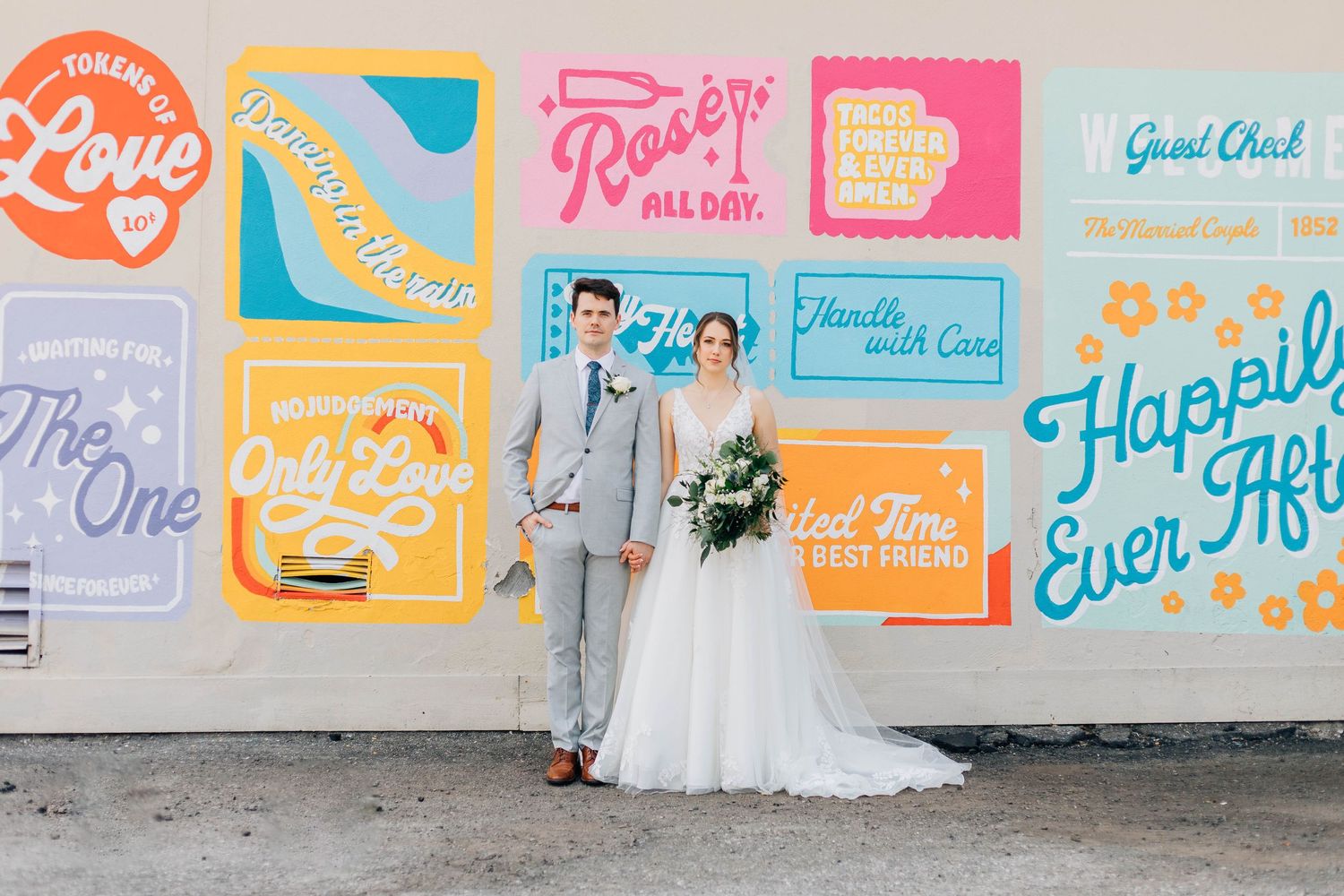 Bride and groom stand before colorful mural wall with wedding-themed typography art.