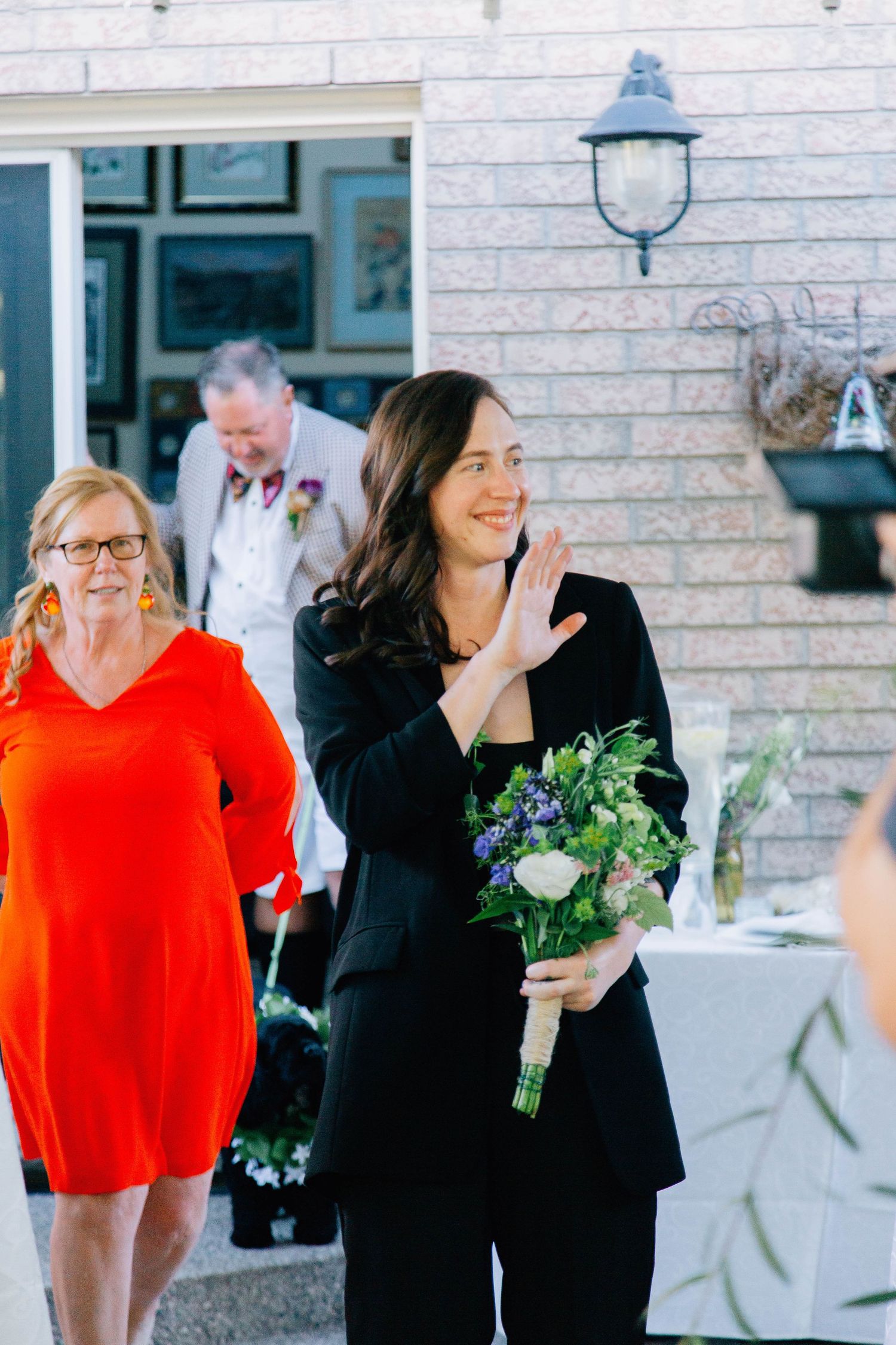 Dark-haired bride in black suit waves while walking down the aisle carrying a blue and white bouquet at backyard wedding.