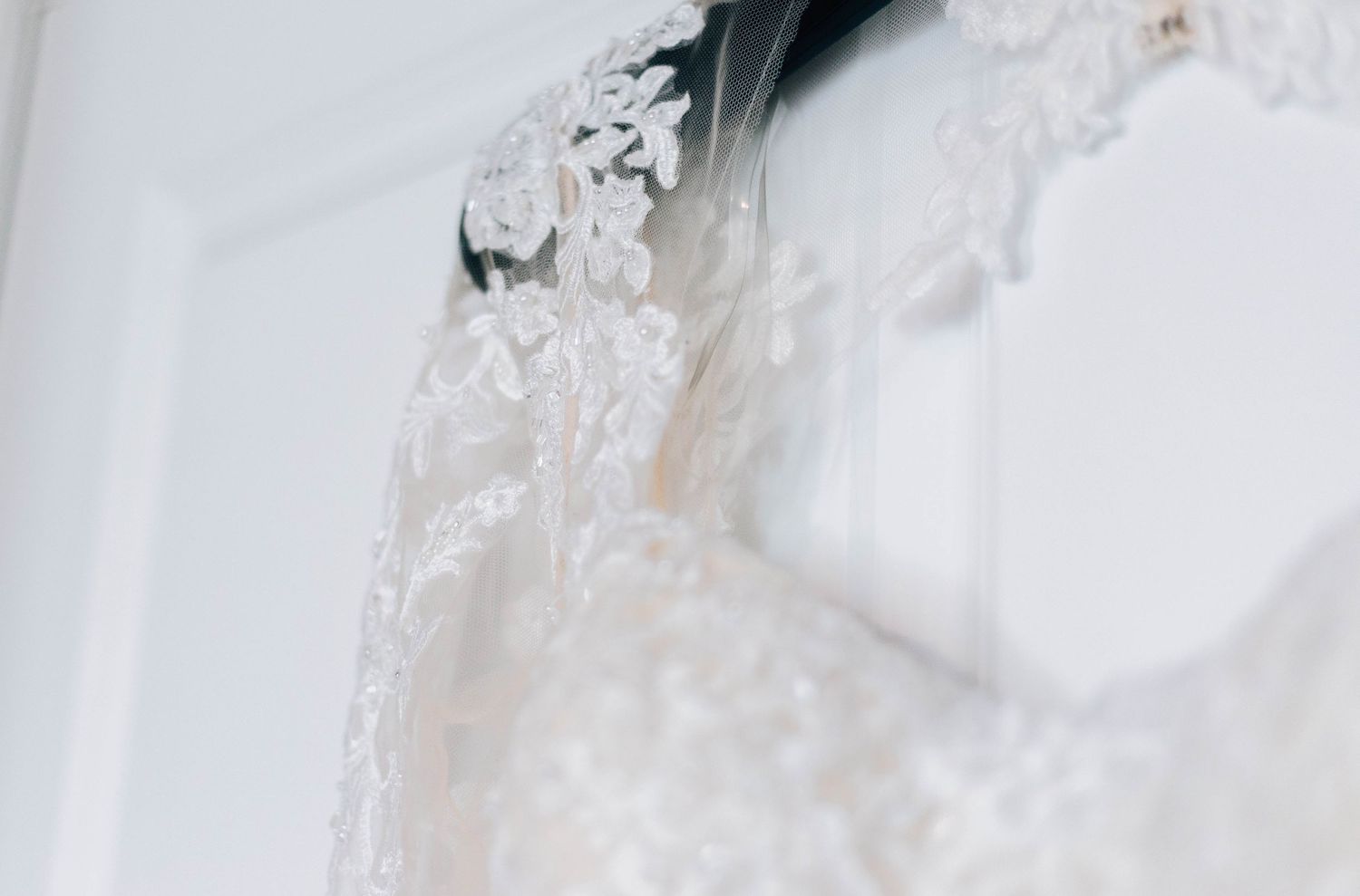 Close-up detail of elegant lace bridal gown hanging on white door.