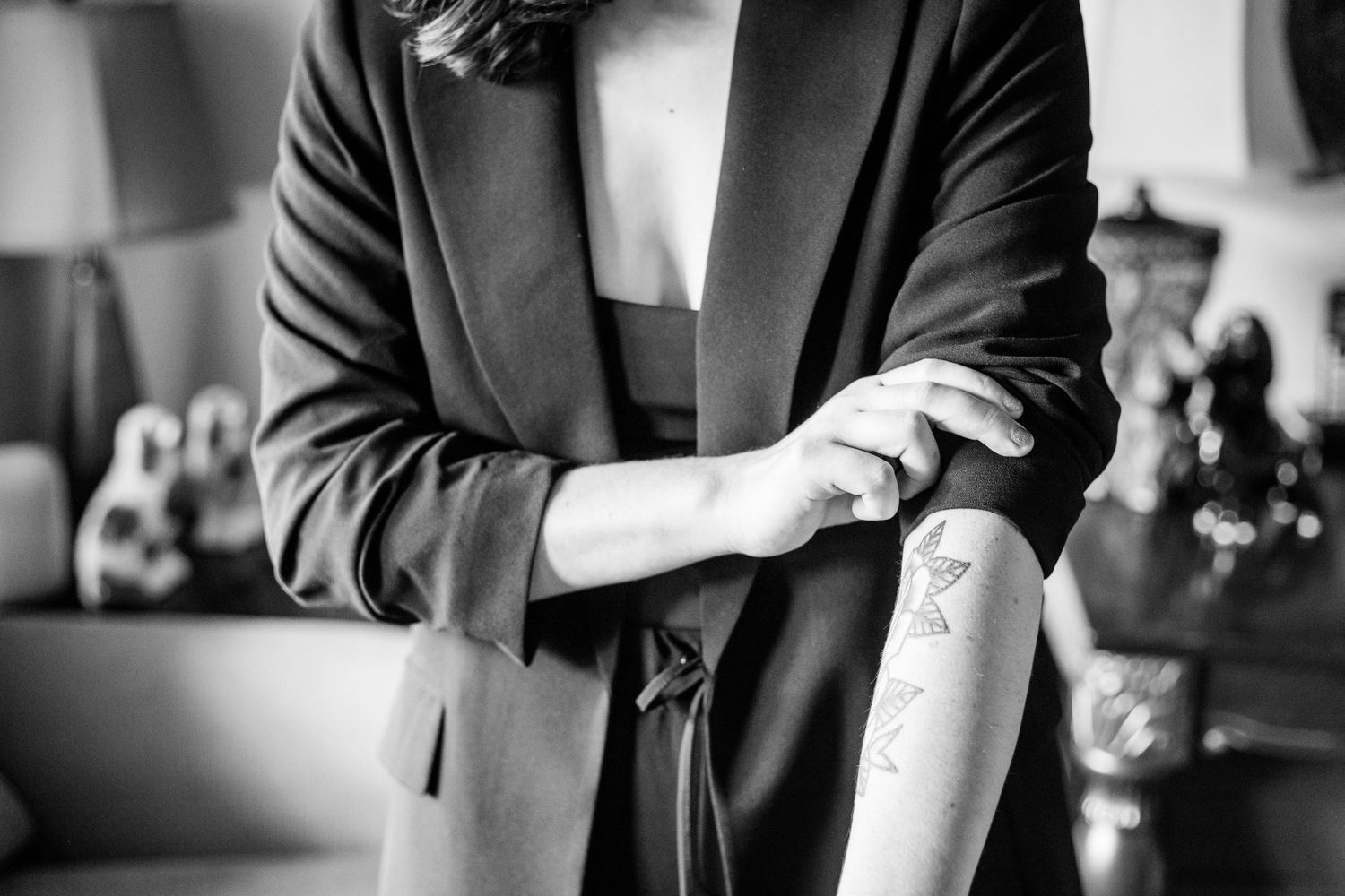 Black and white close-up of person in blazer adjusting sleeve revealing forearm tattoo.