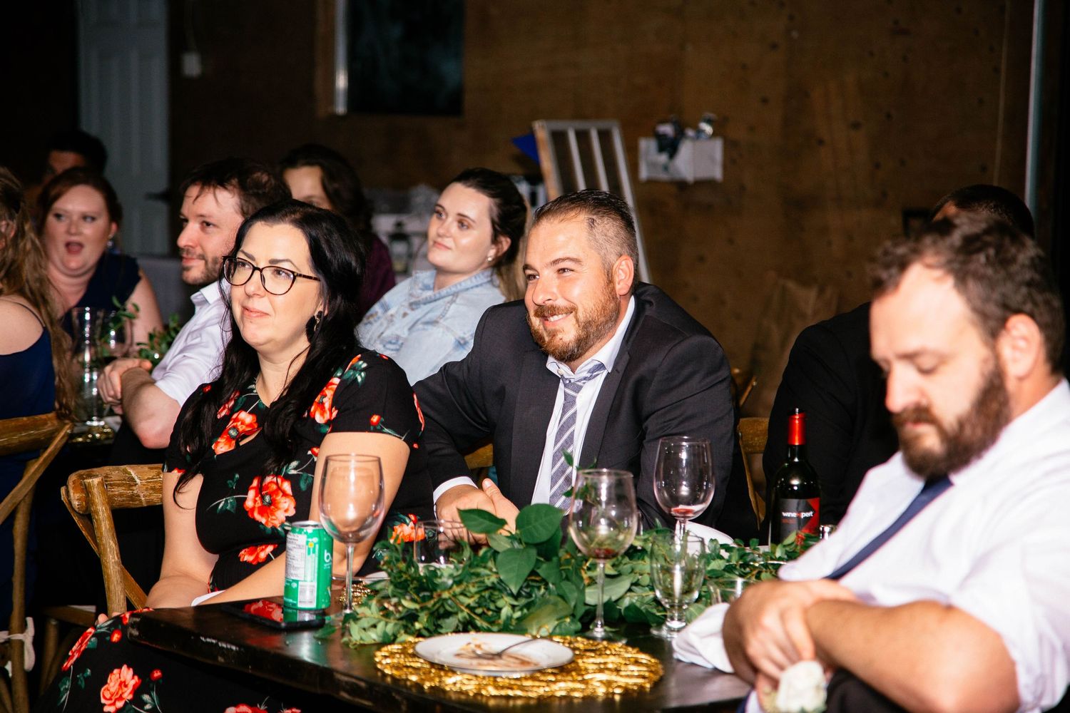 Wedding guests seated at decorated reception tables with greenery centerpieces and wine glasses.