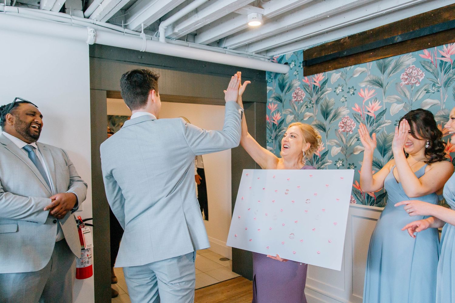 Groom and bridesmaid in lavender dress celebrate with high five while guests applaud at wedding reception.