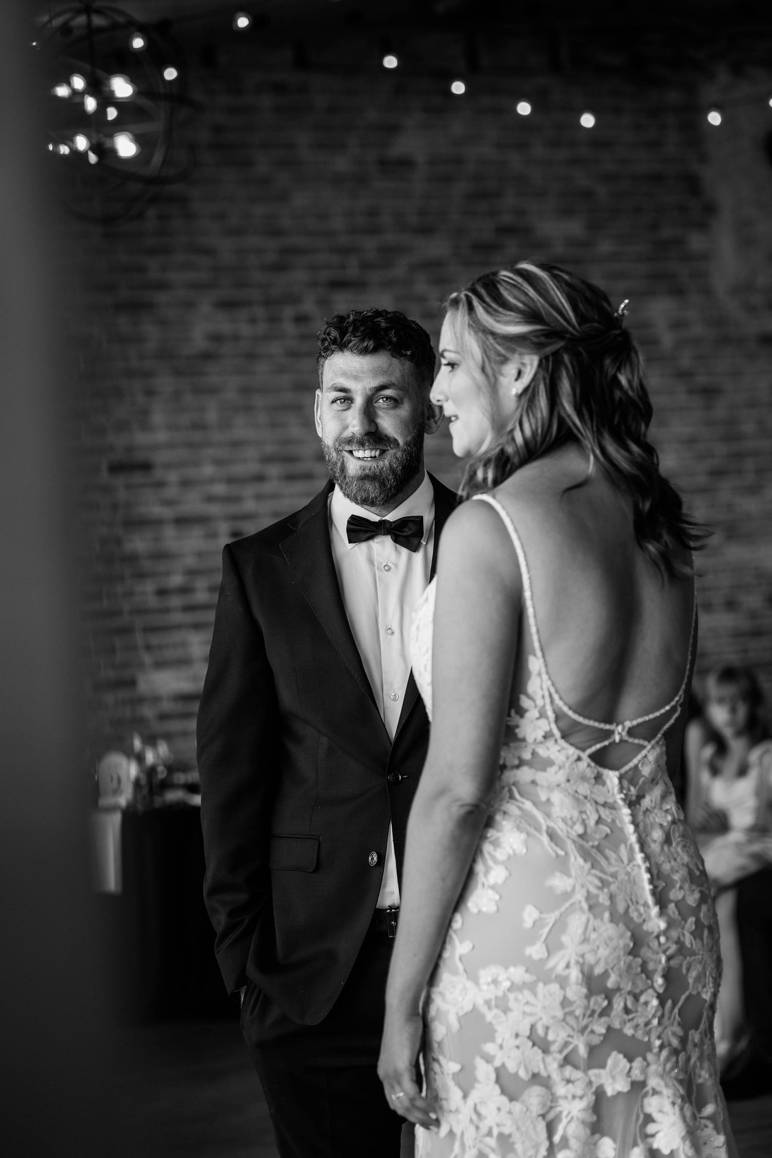 Groom in tuxedo smiling at bride during first dance in black and white photo.