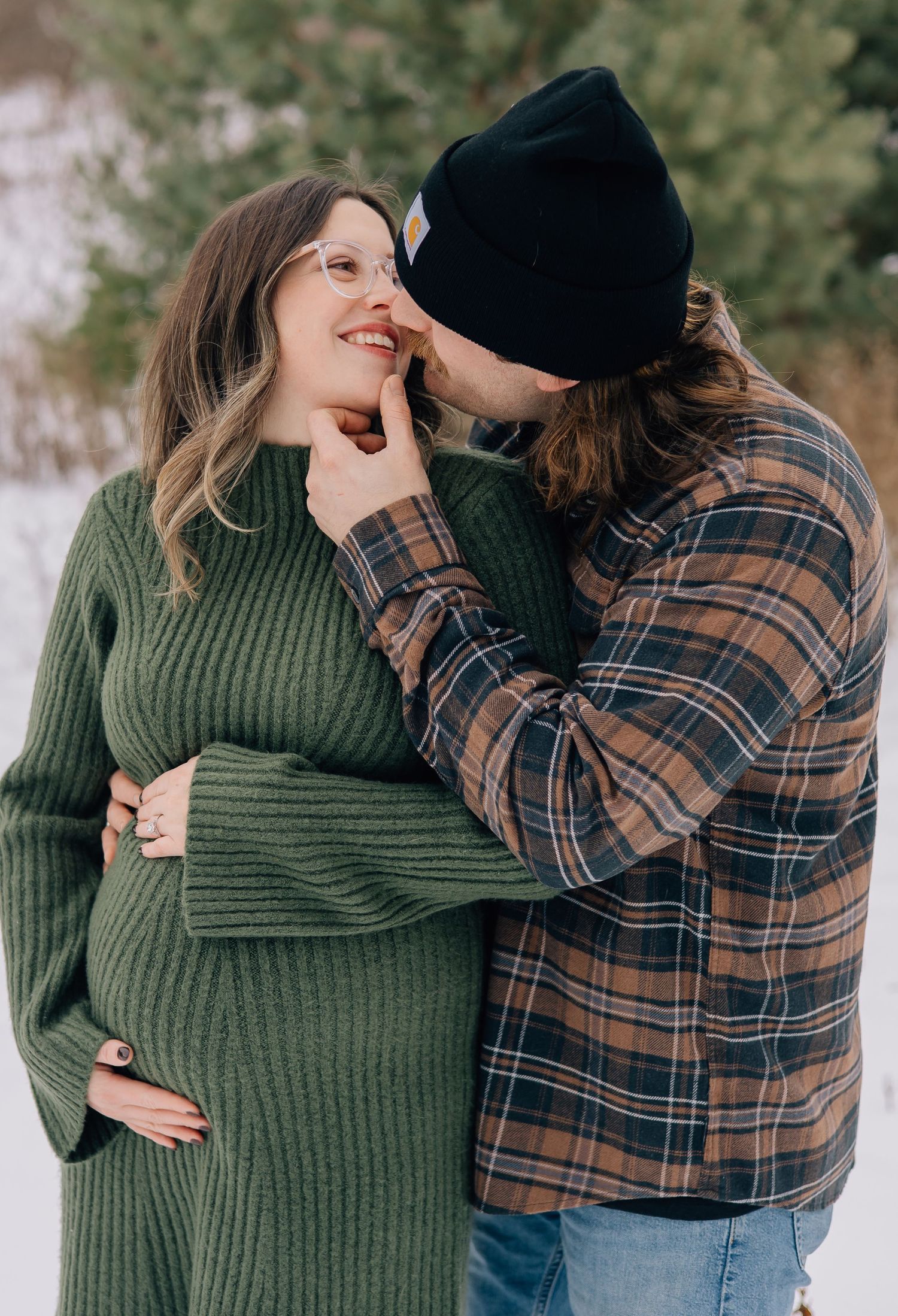 Pregnant couple sharing tender moment outdoors in winter, man kissing woman in green maternity sweater.