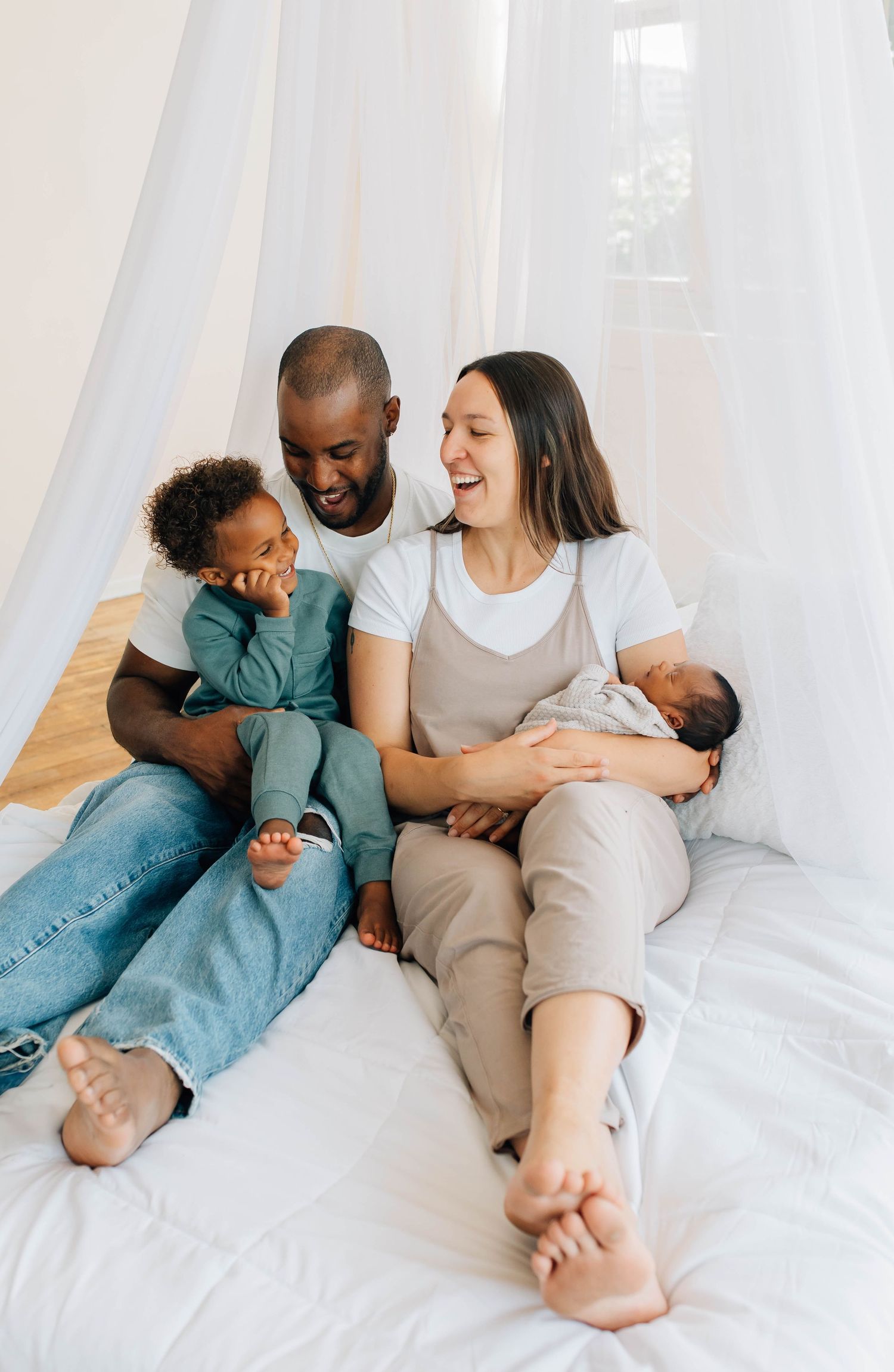 Happy interracial family laughing together on white bed under canopy with toddler and newborn baby.