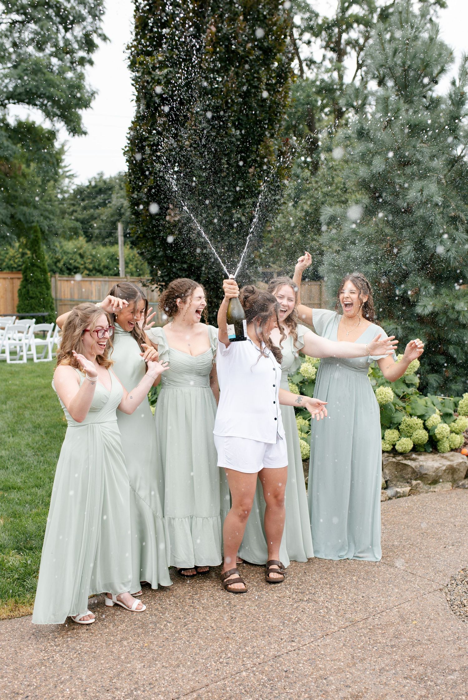 Bride in white sprays champagne with bridesmaids in sage green dresses celebrating outdoors at a garden wedding.