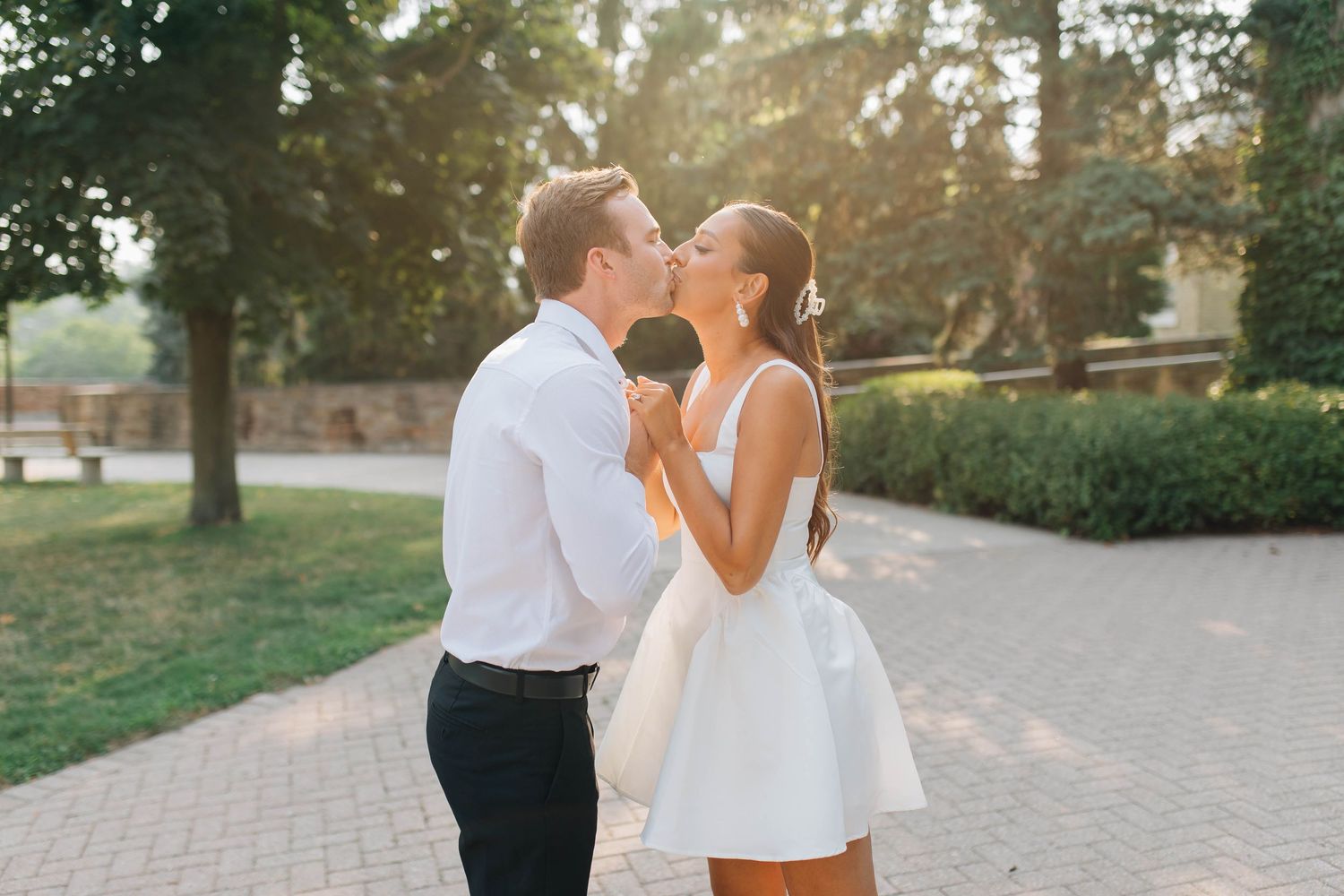 Bride and groom sharing a romantic kiss outdoors in warm golden sunlight with green trees in the background.