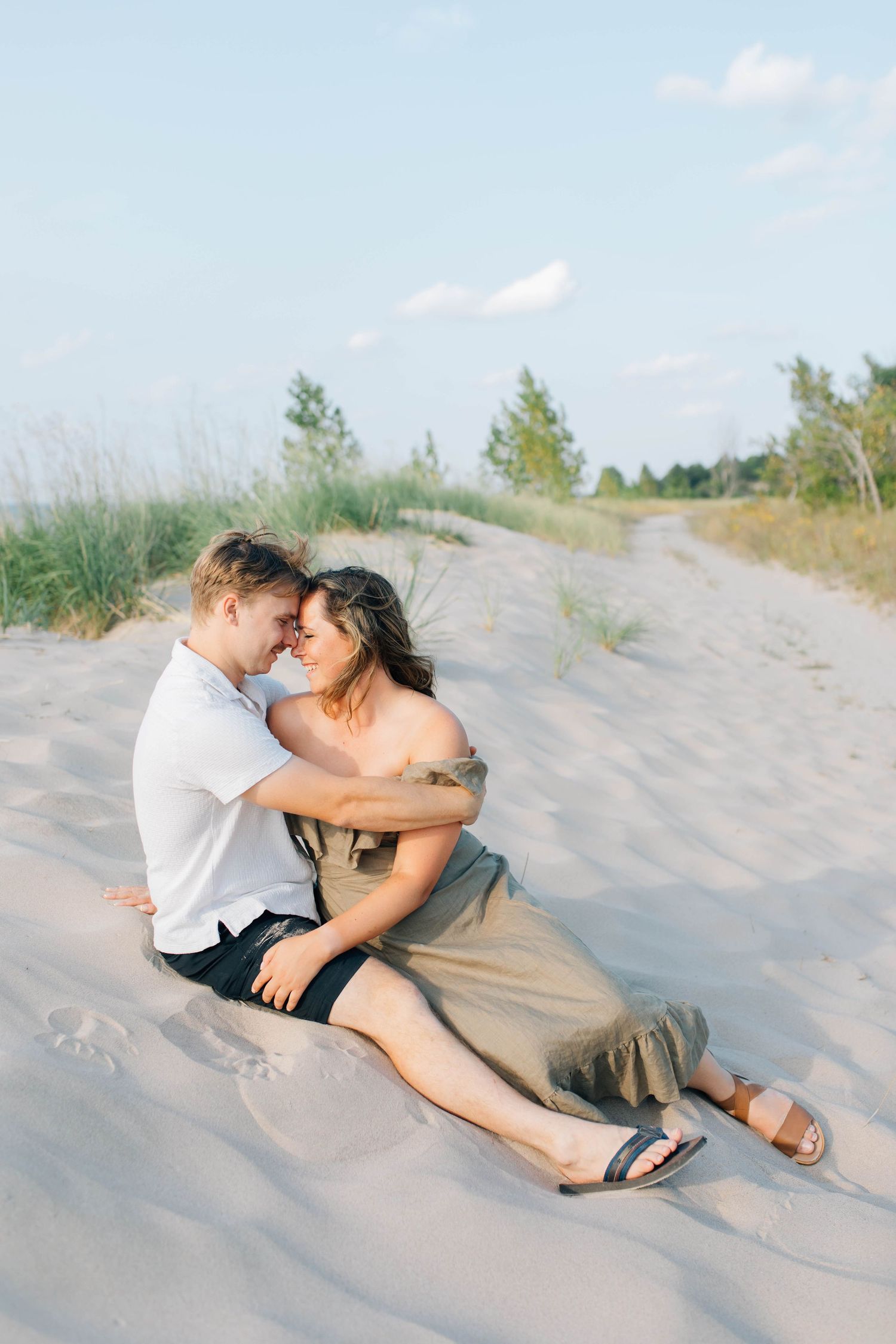 Romantic couple embracing on sandy beach dunes, woman in olive dress and man in white shirt at sunset.