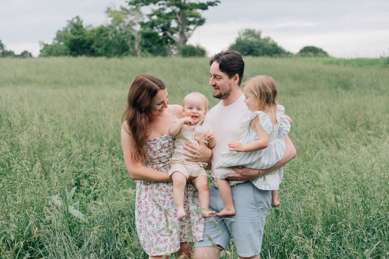 Happy family of four with two young children posing together in a lush green meadow.