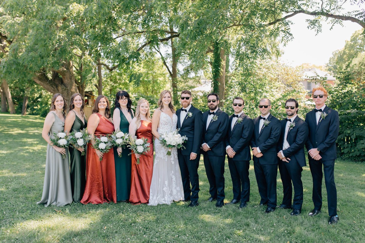 Wedding party posing on green lawn, groomsmen in black tuxedos and bridesmaids in earth tones.