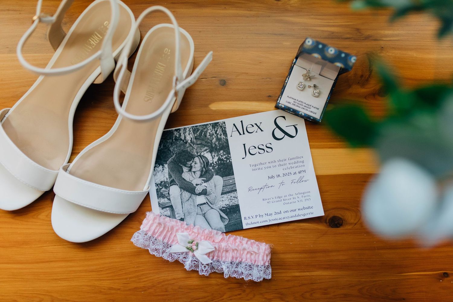 Alex and Jess wedding invitation with white heels, garter, and jewelry box on wooden table.