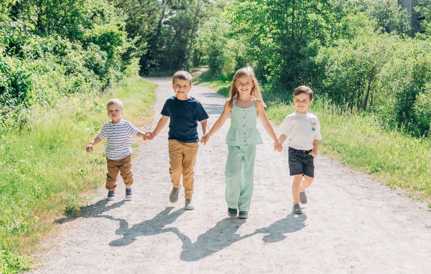 Four happy children running hand-in-hand down a bright, green tree-lined pathway.