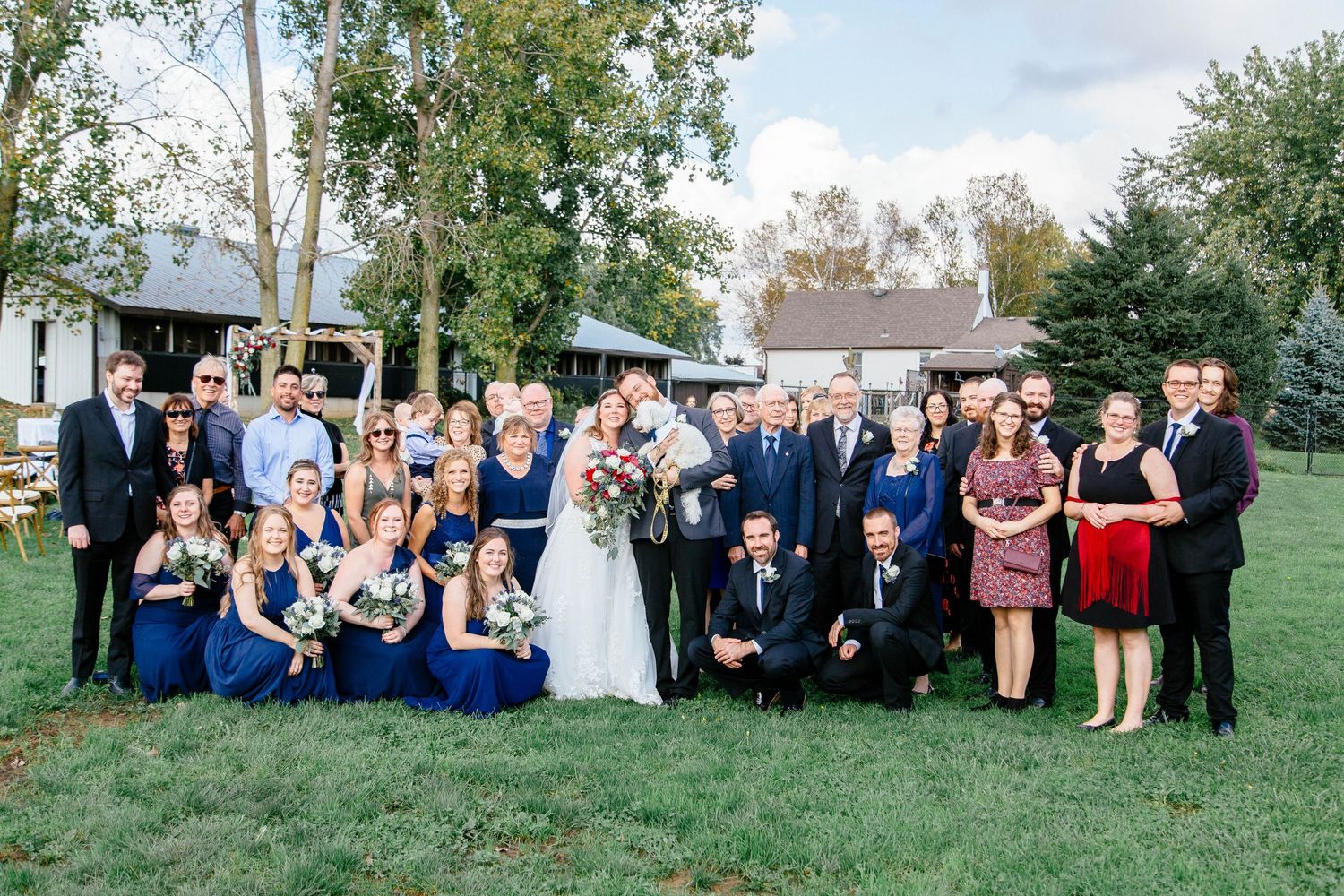 Large wedding party poses together on green lawn at outdoor farm venue on autumn wedding day.