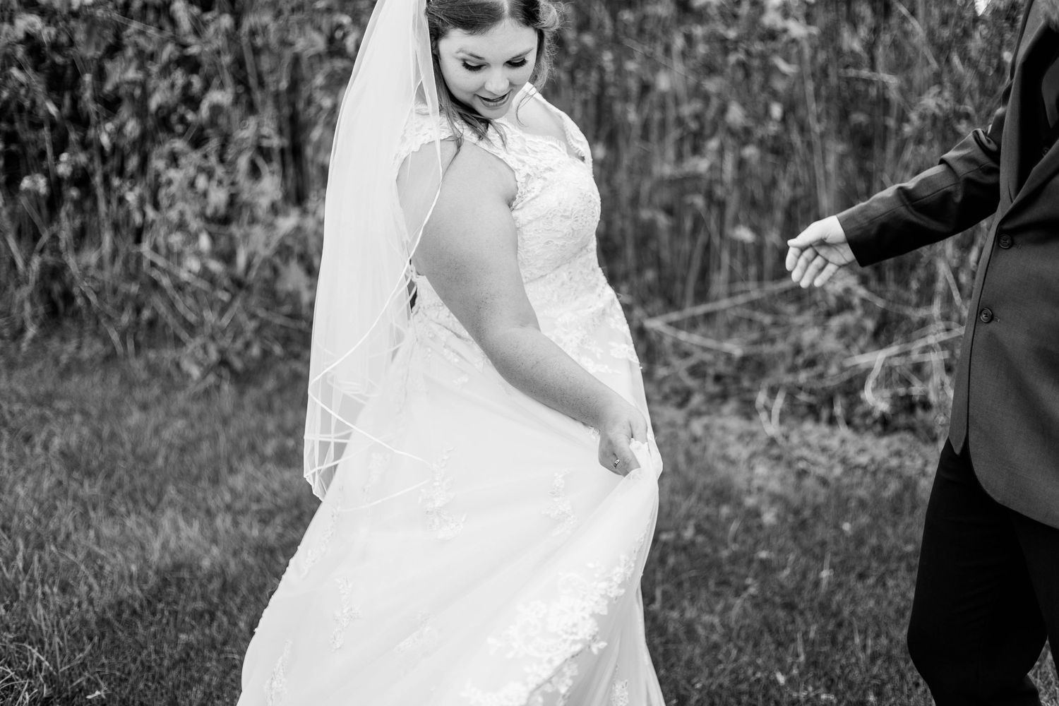 Black and white photo of bride in lace gown lifting dress while walking with groom.