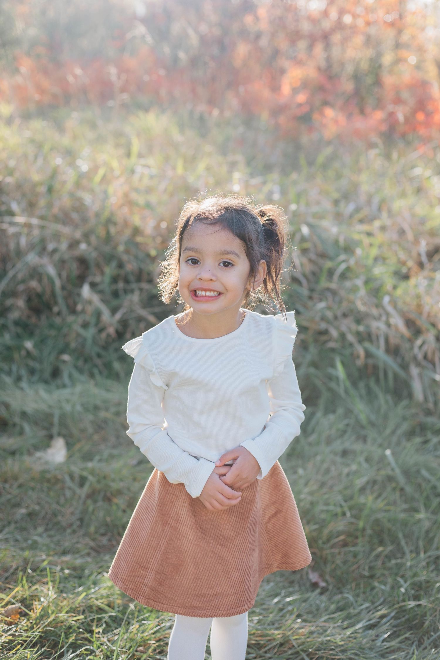 Smiling young girl in white ruffled top and brown skirt standing in sunny autumn field.