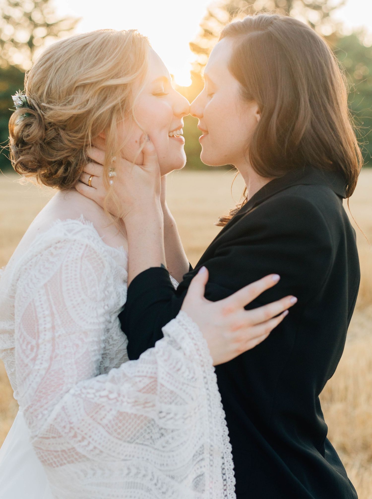 Two brides sharing a romantic sunset kiss in golden field, one in white lace dress, one in black suit.