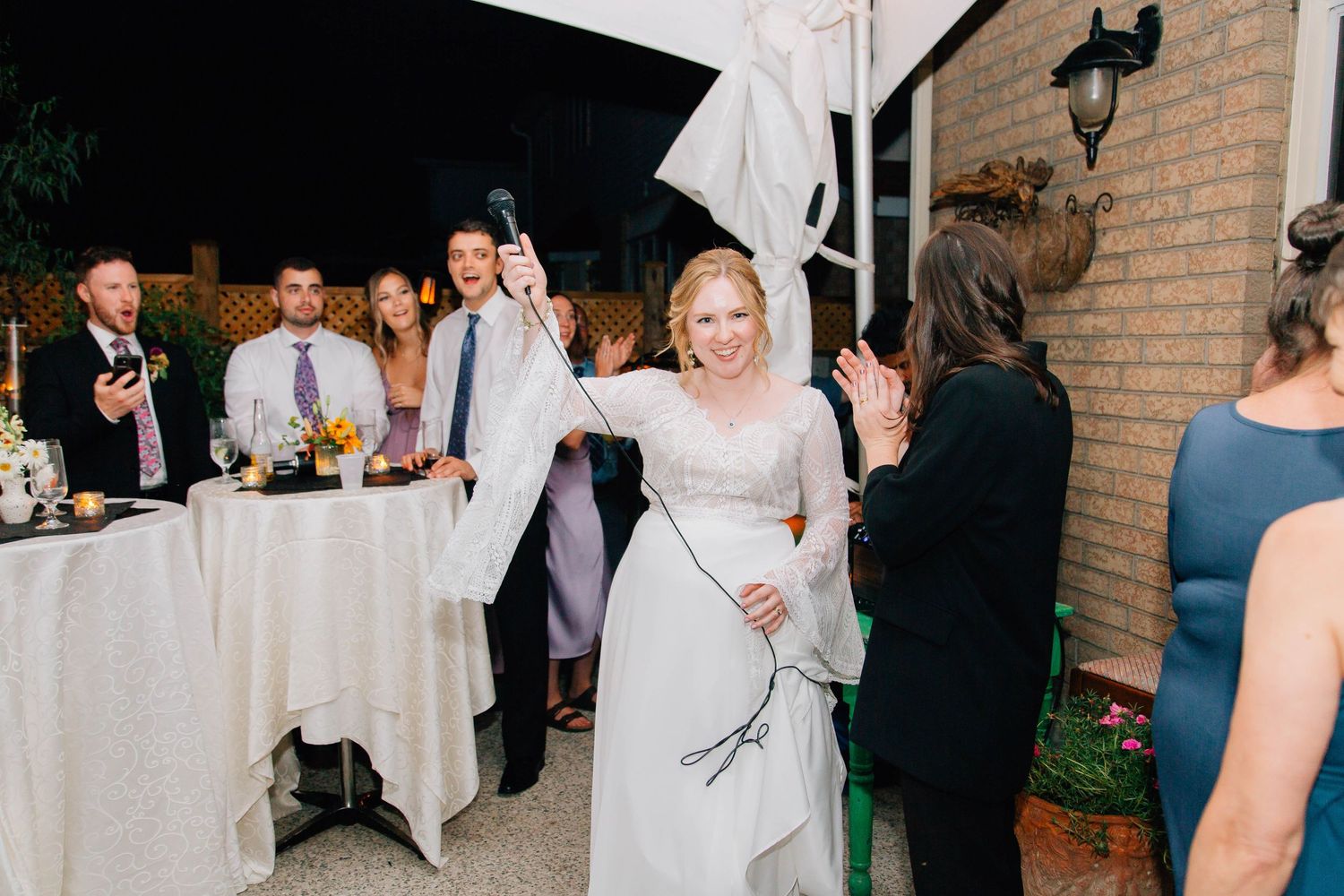Bride in white lace gown walking through cheering wedding guests at outdoor reception.