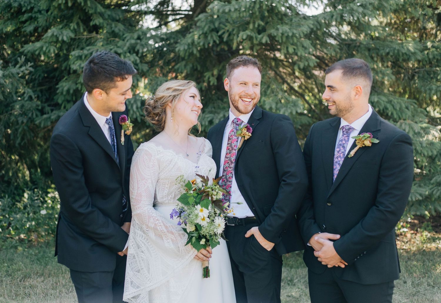 Bride laughing with three groomsmen in black suits standing together near green trees outdoors.
